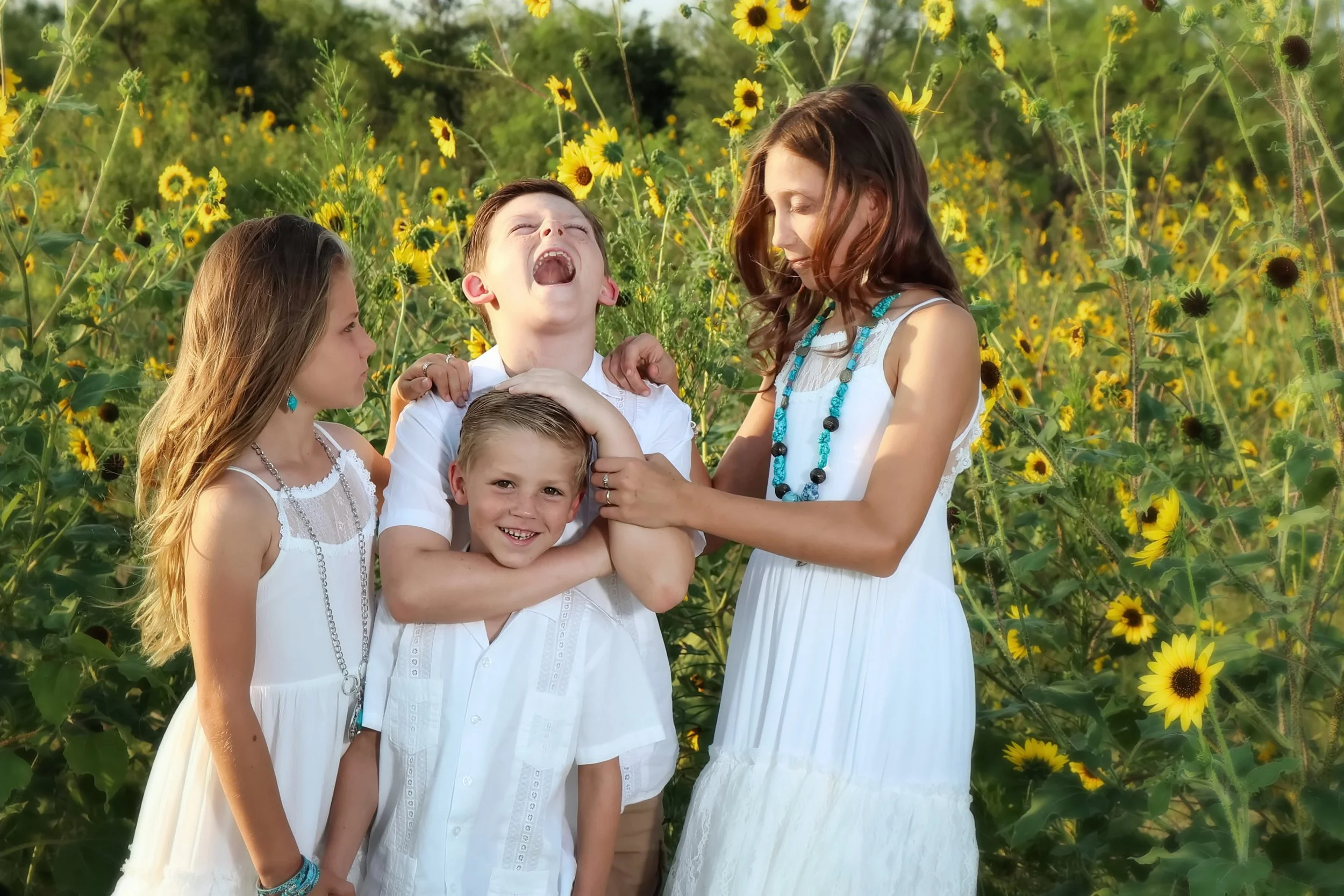 Four children, two boys and two girls, standing in a field of tall yellow sunflowers. The children are dressed in white and are engaging in playful, humorous poses, with one boy laughing and the others making funny faces in Belton, Texas.