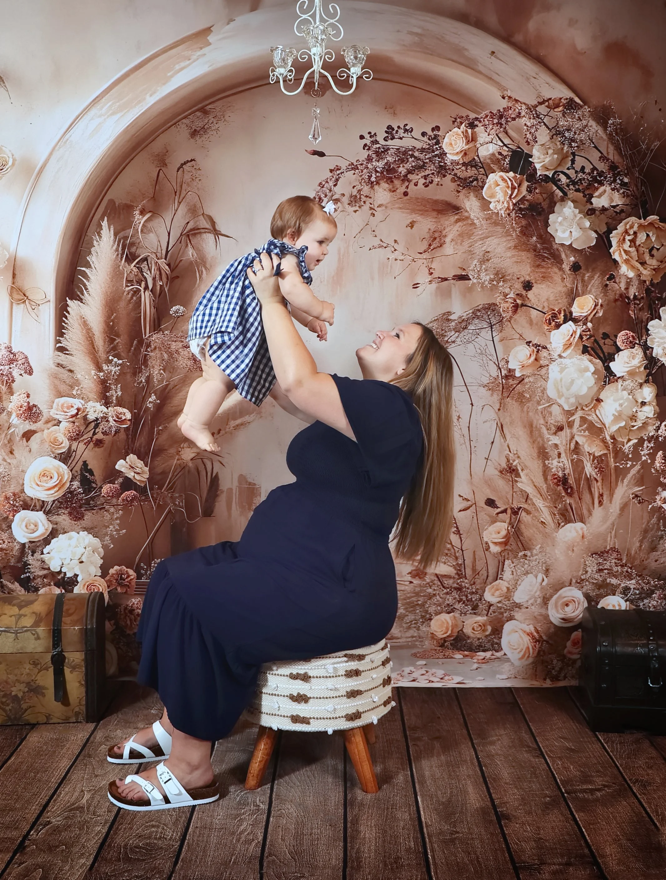 A woman with long hair wearing a navy blue dress is smiling as she lifts a young girl in a blue and white checkered dress in a decorated room with flowers and a chandelier.