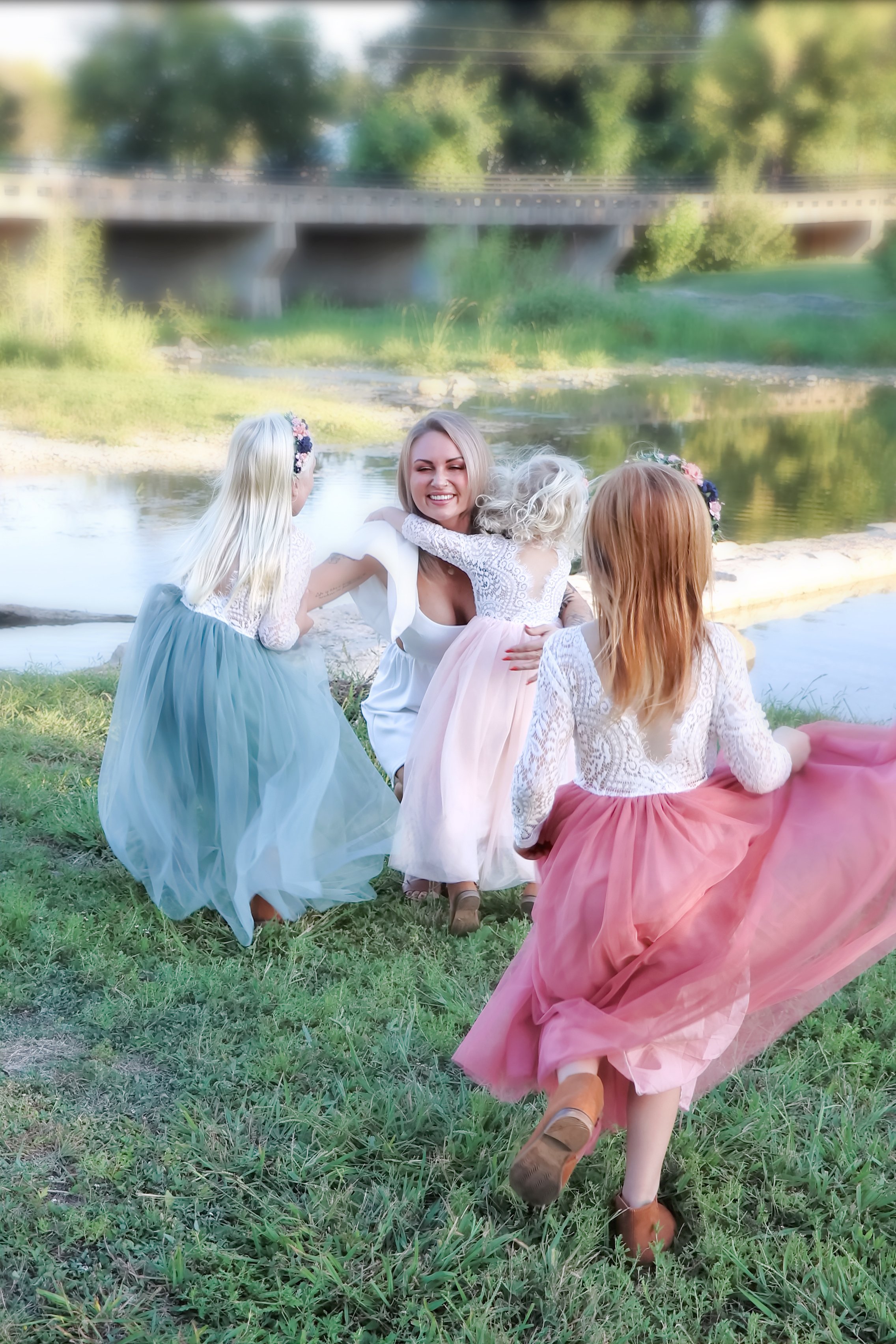 A woman and three young girls in pastel dresses playing by a river, with a bridge and trees in the background in Salado, Texas.