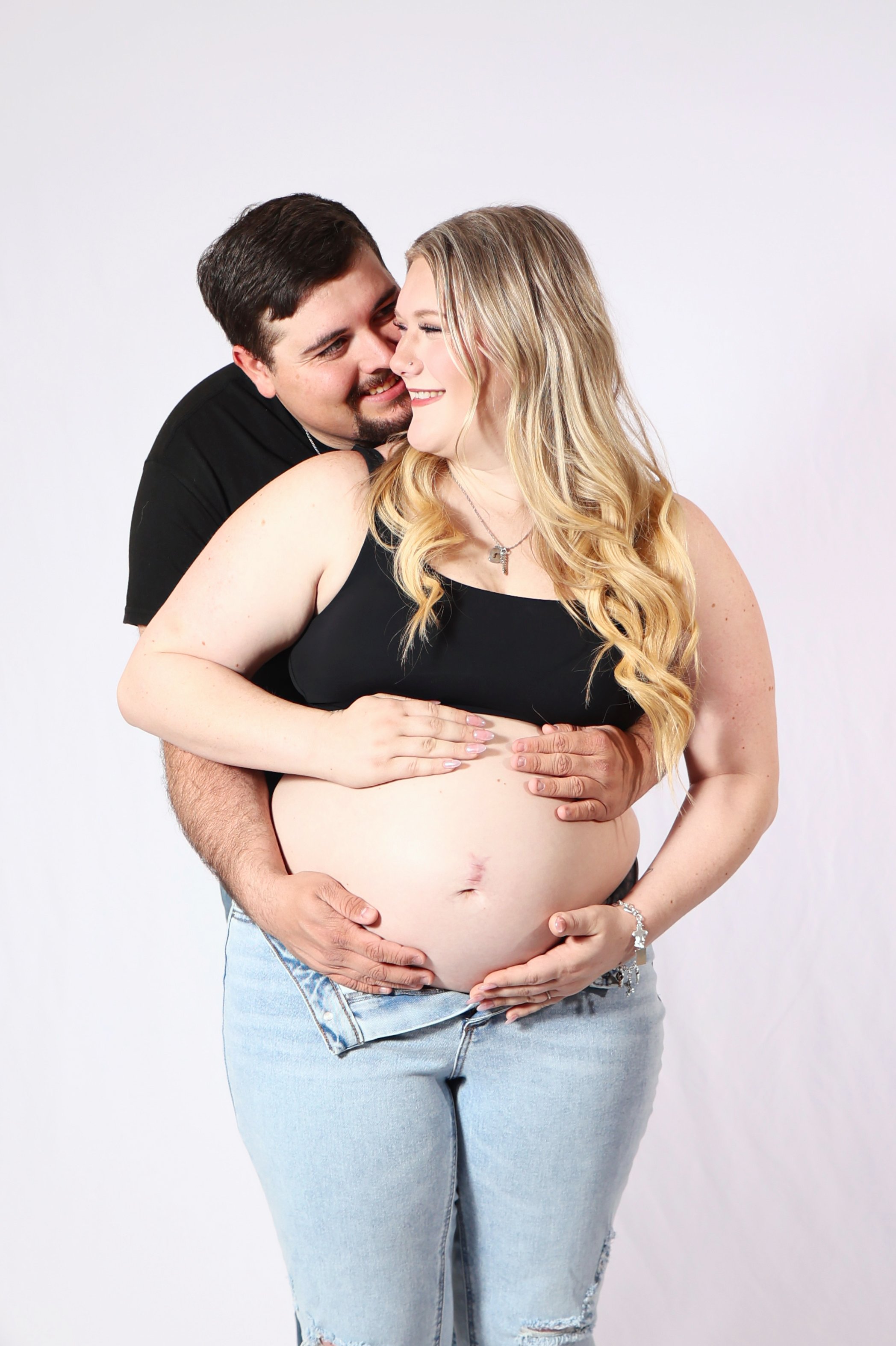 A pregnant woman and her partner sharing a joyful moment, with the partner embracing her from behind and both smiling at each other in a Studio Maternity Session
