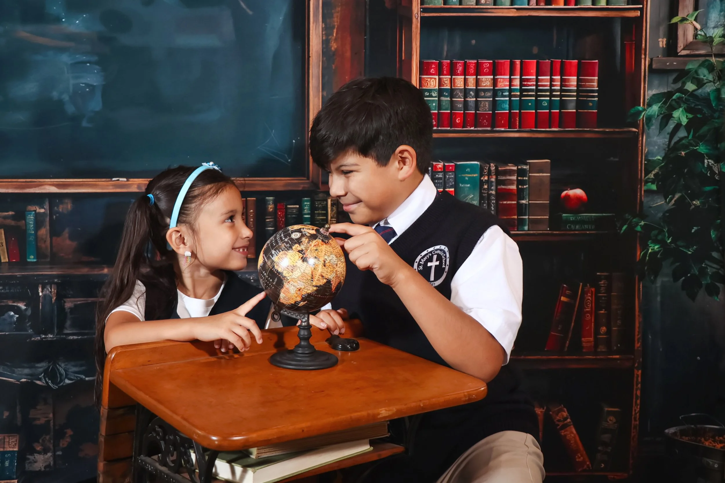 A young brother and sister in school uniforms sitting at a desk, smiling and pointing at a globe, in a room with a bookshelf filled with books and green plants in the background in back to school pictures..