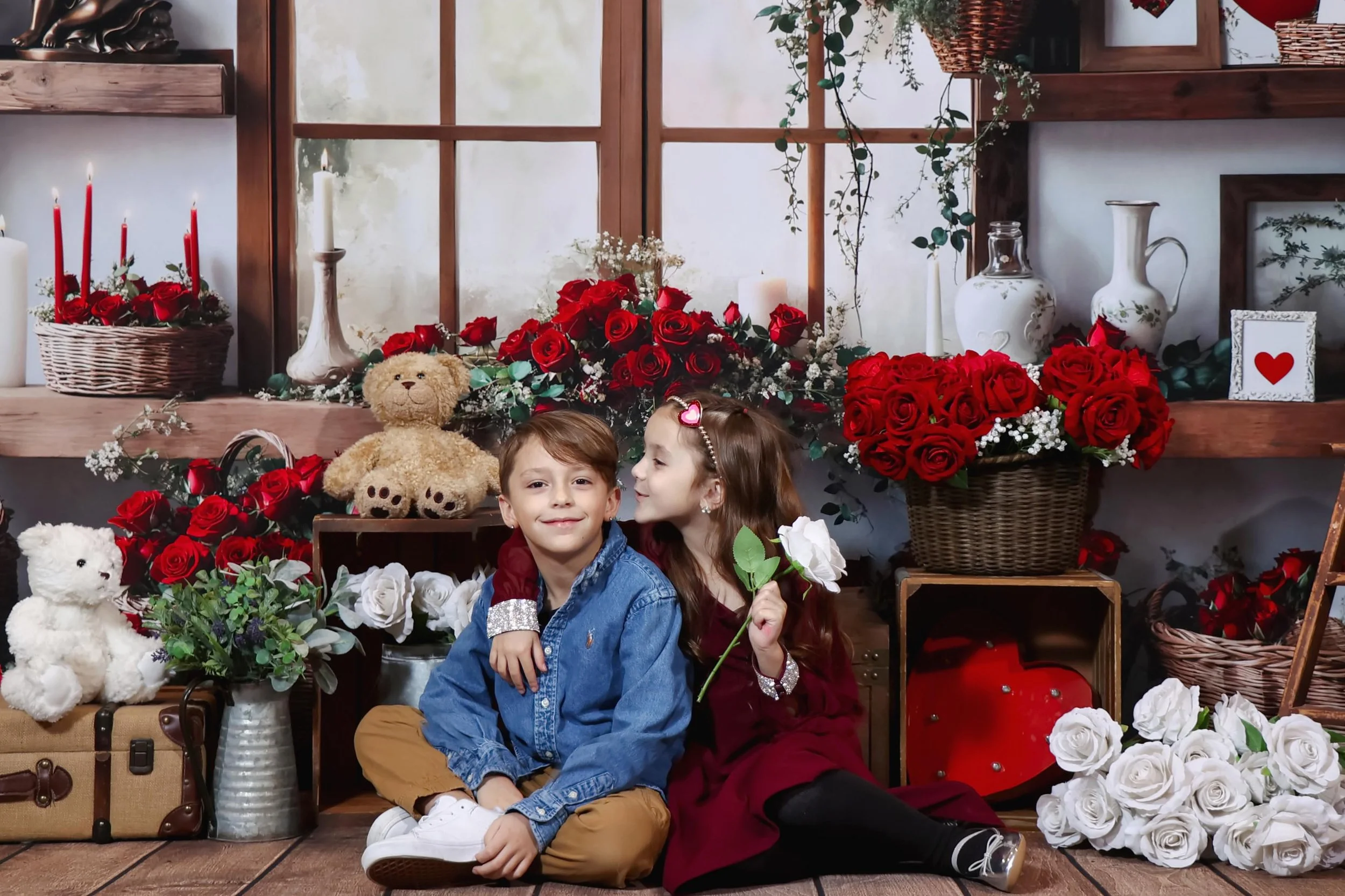 Two children, a boy and a girl, sitting inside a decorated room with flowers and teddy bears, sharing a moment of affection, with the girl holding a flower and the boy smiling.