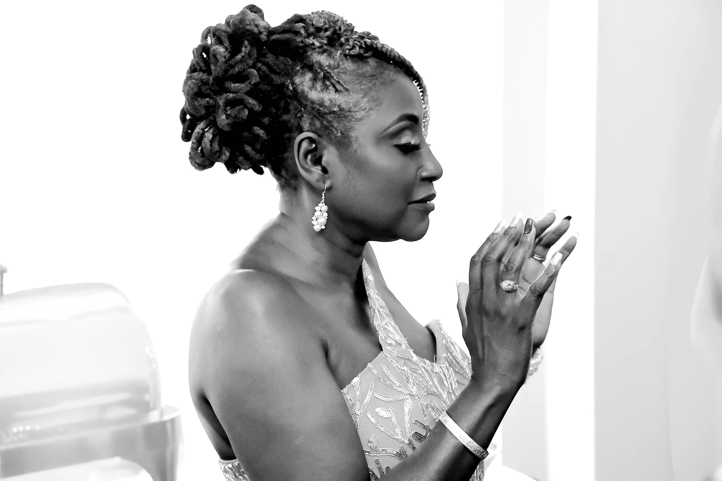 Black woman with styled hair and earrings, praying with eyes closed, hands pressed together, wearing a dress with embroidery, in a white room before her wedding vow renewal ceramony in Temple, Texas.