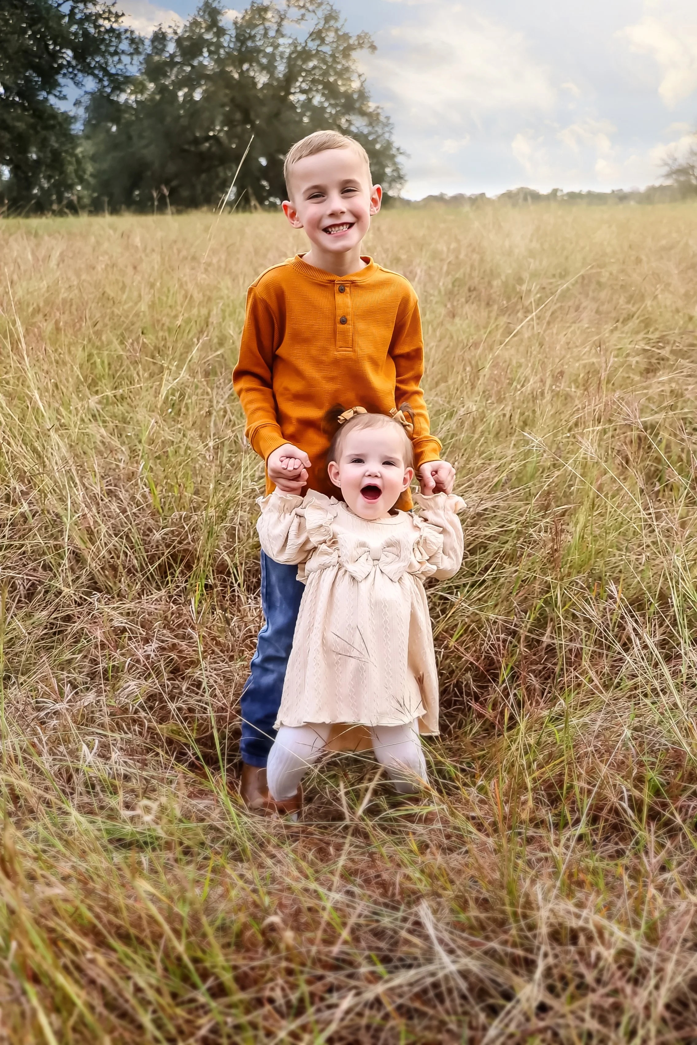 A young boy with short hair wearing an orange top holding a toddler girl in a cream-colored dress, standing in a grassy field at Lake Belton