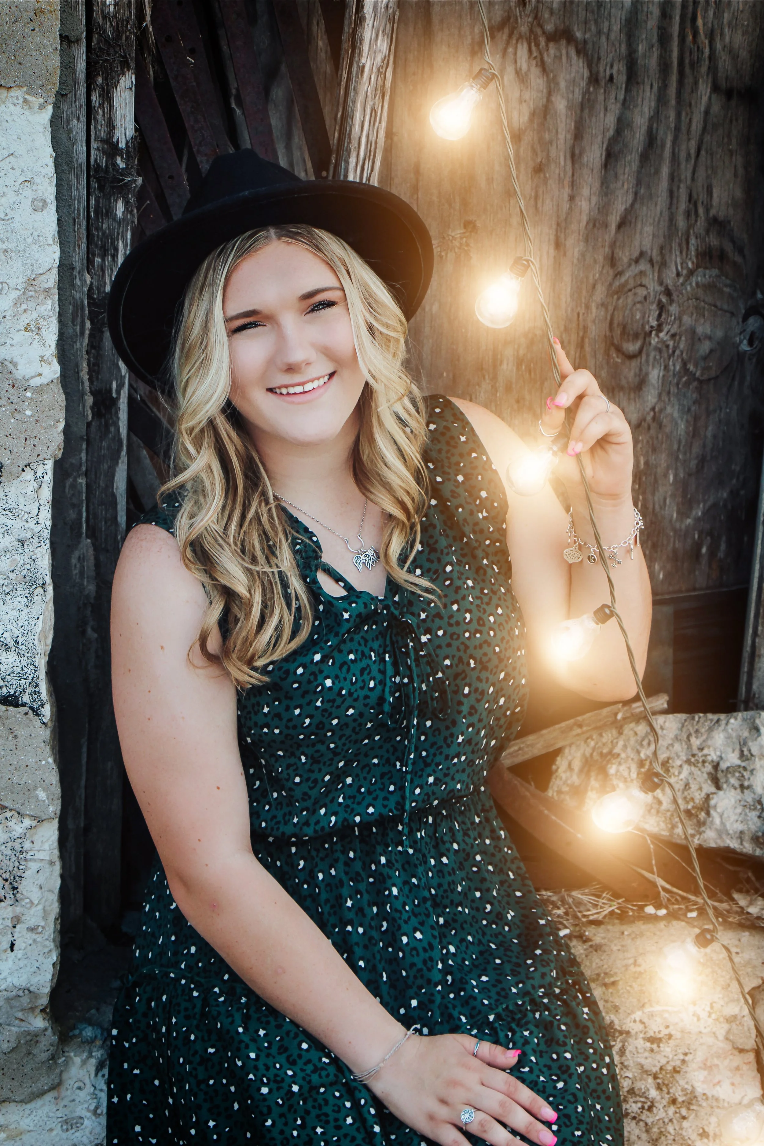 A Graduating Senior with blonde hair wearing a black wide-brimmed hat and a dark green leopard print dress, smiling while holding a string of glowing lights, standing against a rustic wooden and stone background in Downtown Belton..