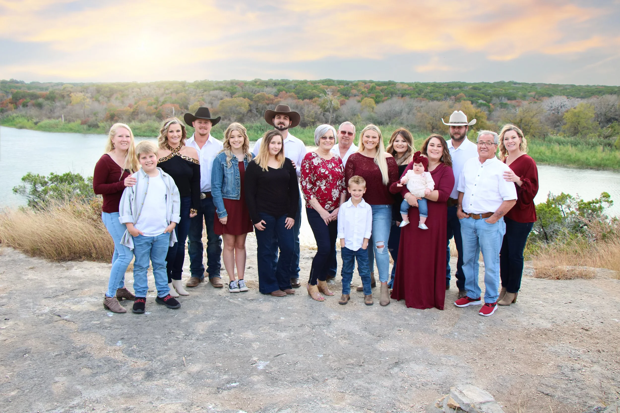 A large family group posing outdoors by a river at sunset with trees in the background. The group includes adults and children, some wearing casual clothing and others dressed more formal or country style, with a few wearing cowboy hats in Clifton.