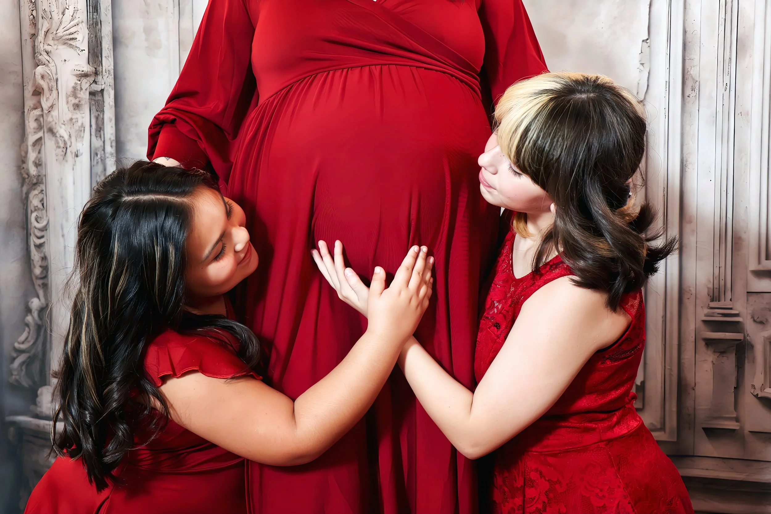 Two young girls in red dresses gently touching their pregnant mom's belly, lovingly looking at it in Belton, Texas.
