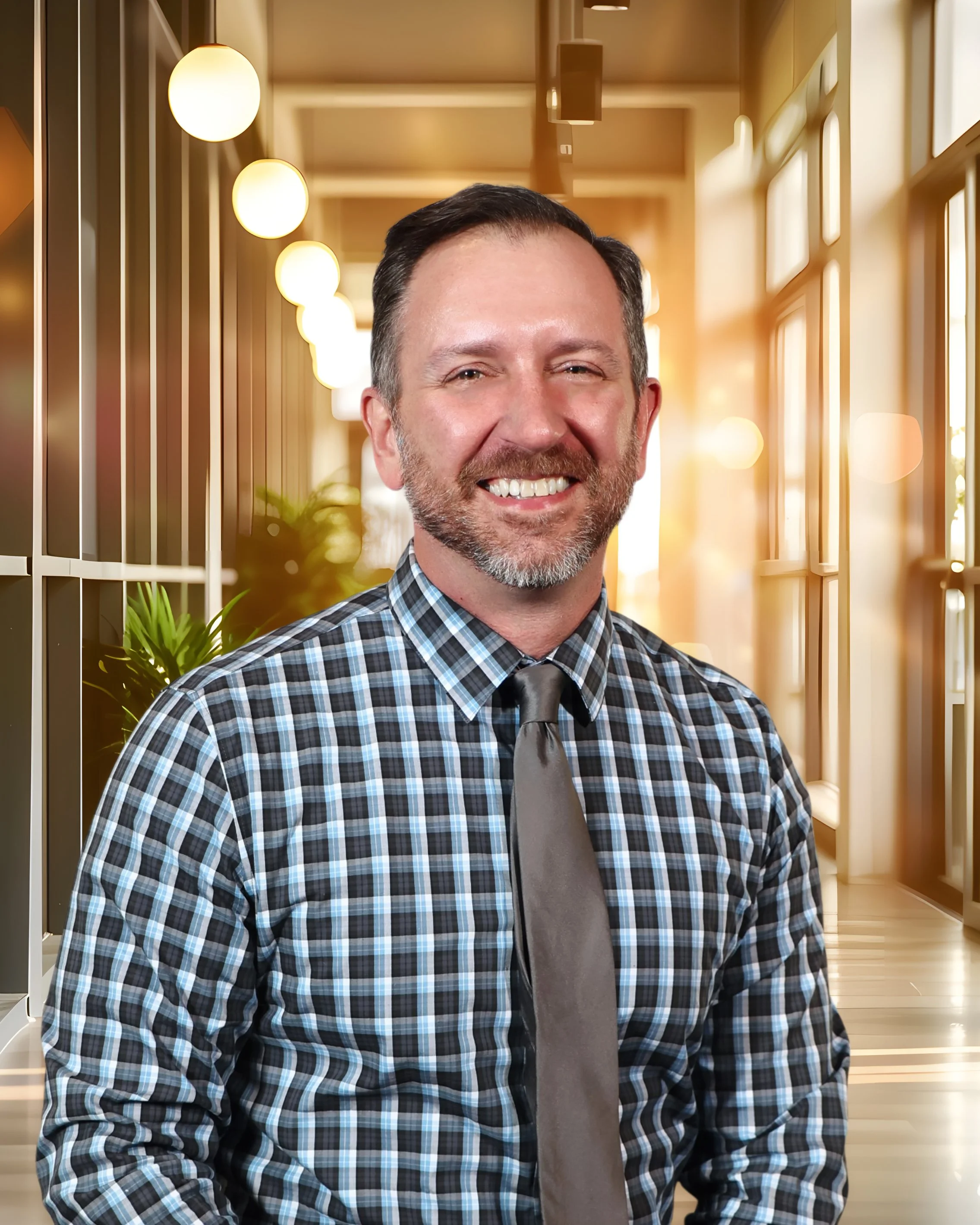 A smiling man with a beard and short dark hair, wearing a plaid shirt and gray tie, standing in a brightly lit indoor space with large windows and decorative lighting.