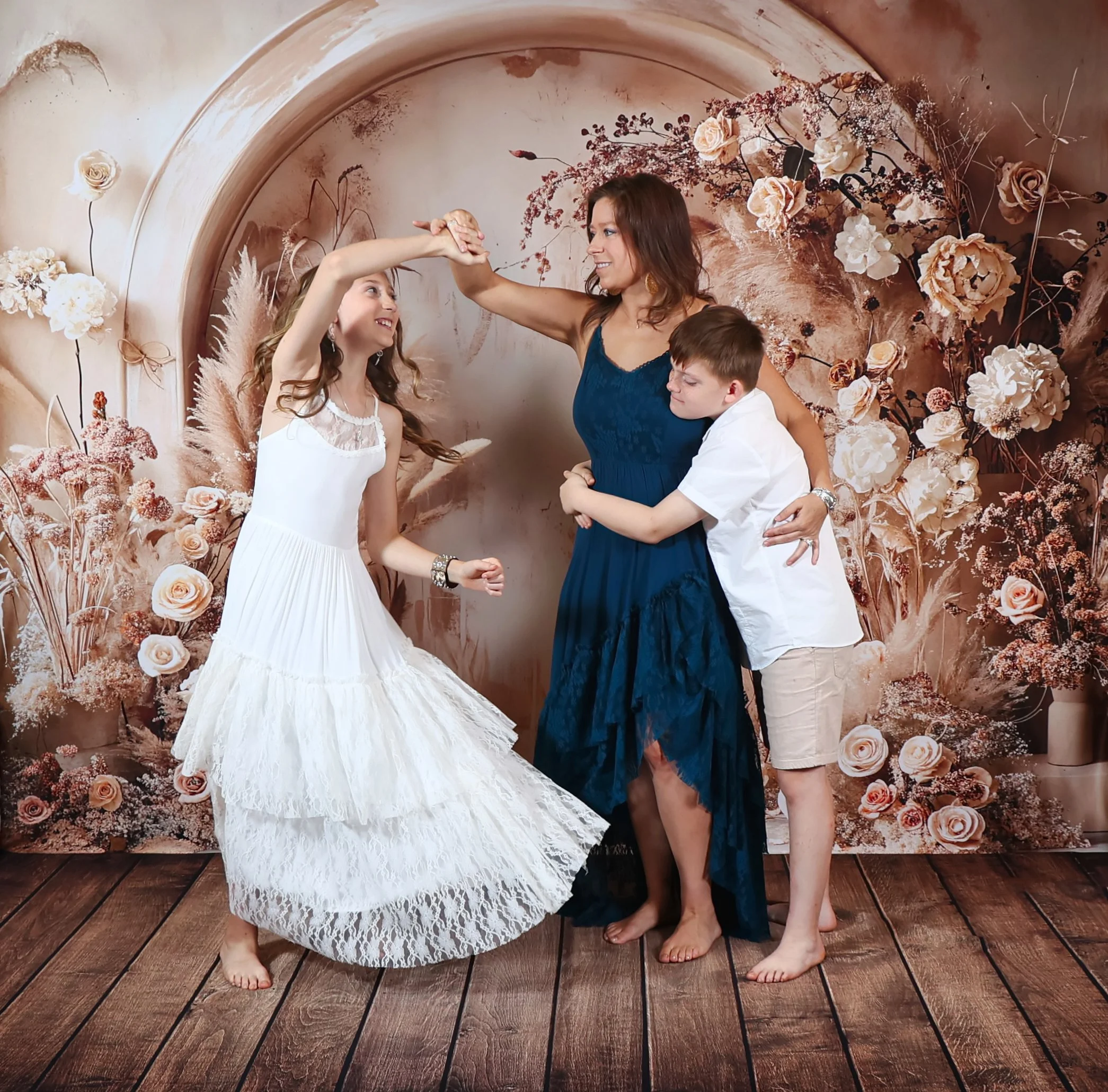 Megan Gibson in a blue dress dancing with a girl in a white dress while a boy in a white shirt and khakis hugs her in front of a floral backdrop.