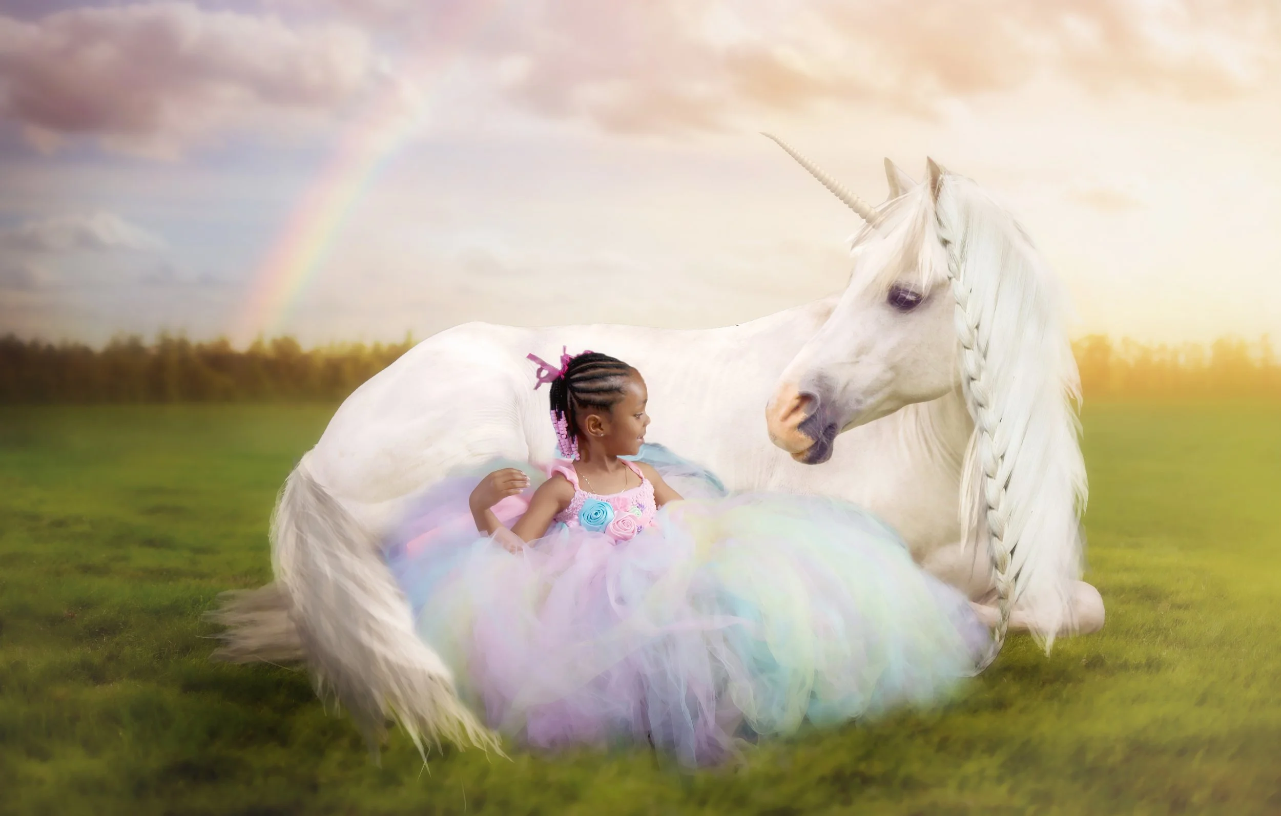 A young girl with braided hair and a colorful dress sitting on grass next to a resting white unicorn with a braided mane, with a rainbow in the cloudy sky in the background.