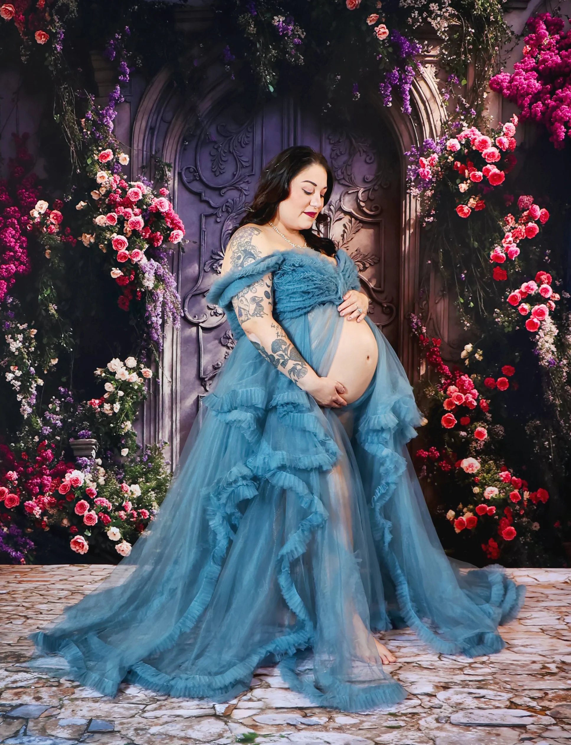 Pregnant woman wearing a flowing blue dress with ruffles, standing on a stone-paved surface in front of a floral backdrop with pink and purple flowers and carved wood details in a Studio Maternity Session.