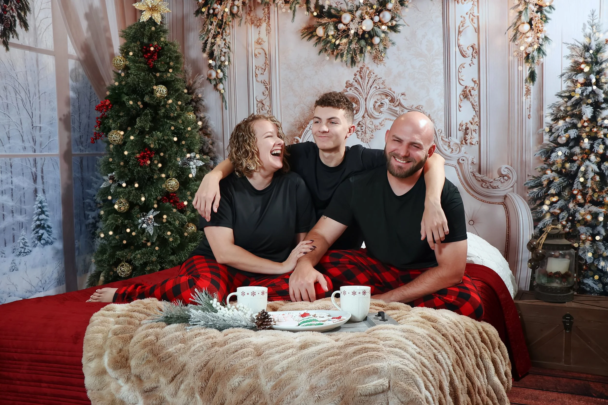 A happy family of three sitting together on a bed in a decorated Christmas room, with Christmas trees, ornaments, and a snowy window in the background, wearing matching black shirts and red plaid pajamas for cozy Christmas pictures at MGP studio