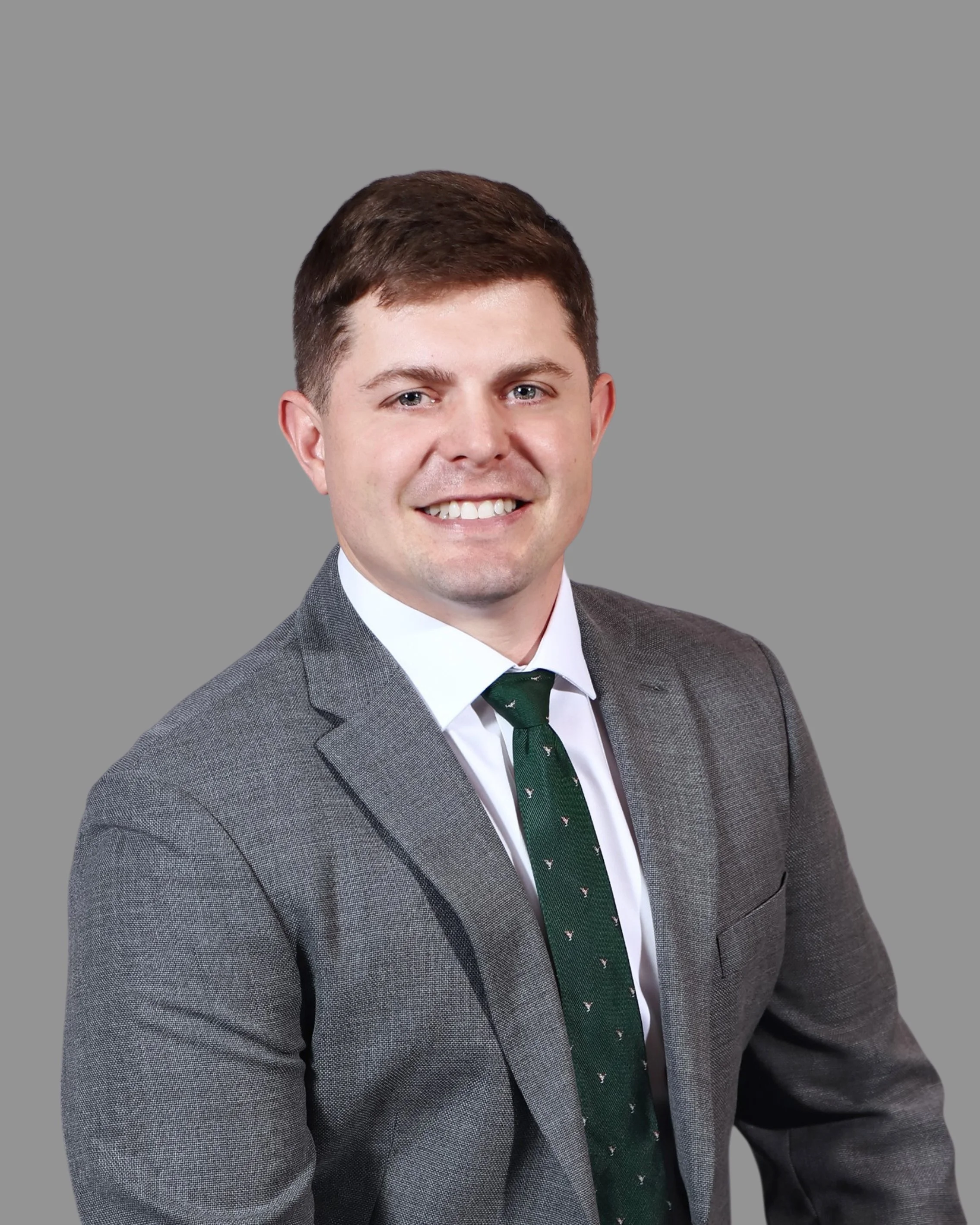 Headshot of a young man with short brown hair smiling, wearing a gray suit, white shirt, and a green tie with small patterns, against a plain gray background.