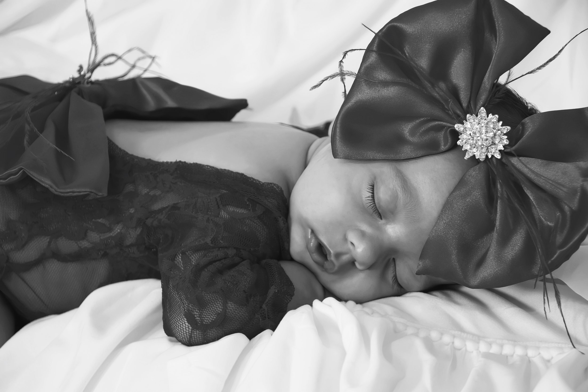 A newborn with a large bow headband, wearing lace black clothing, sleeping on a bed with white pillows in an on location newborn photography session.