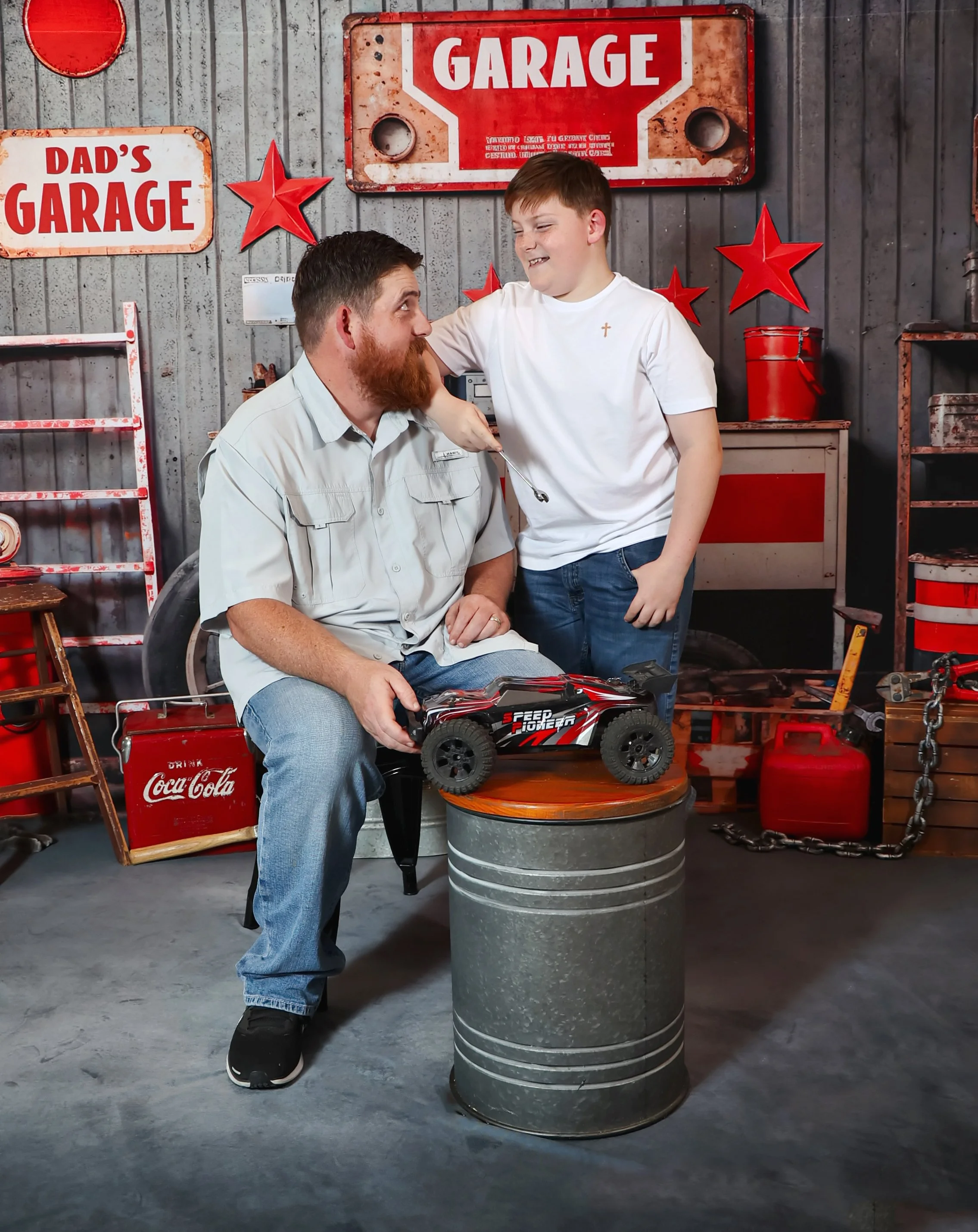 A man with a beard and a boy with short hair in a garage decorated with red stars and vintage signs. The man is seated on a stool, holding a remote-controlled toy car, while the boy stands beside him, smiling and placing his hand on the man's head.