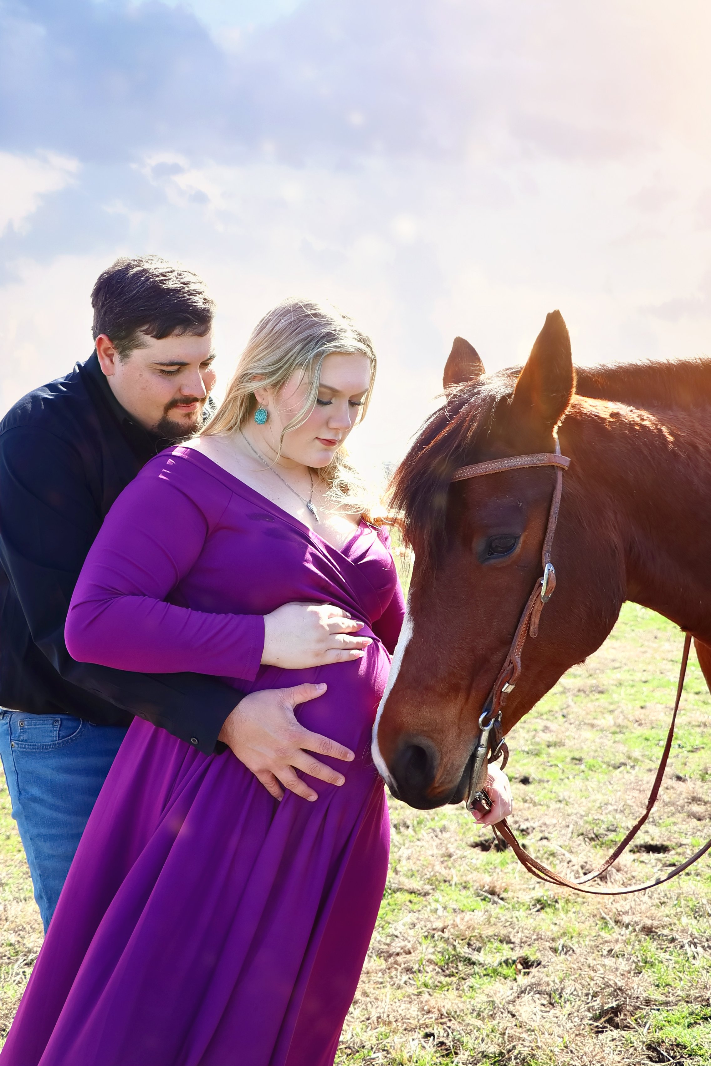 A pregnant woman in a purple dress standing in a field with a brown horse, with a man standing behind her and holding her belly, during daytime with a partly cloudy sky. in an on location Maternity photography session in Clifton, Texas.