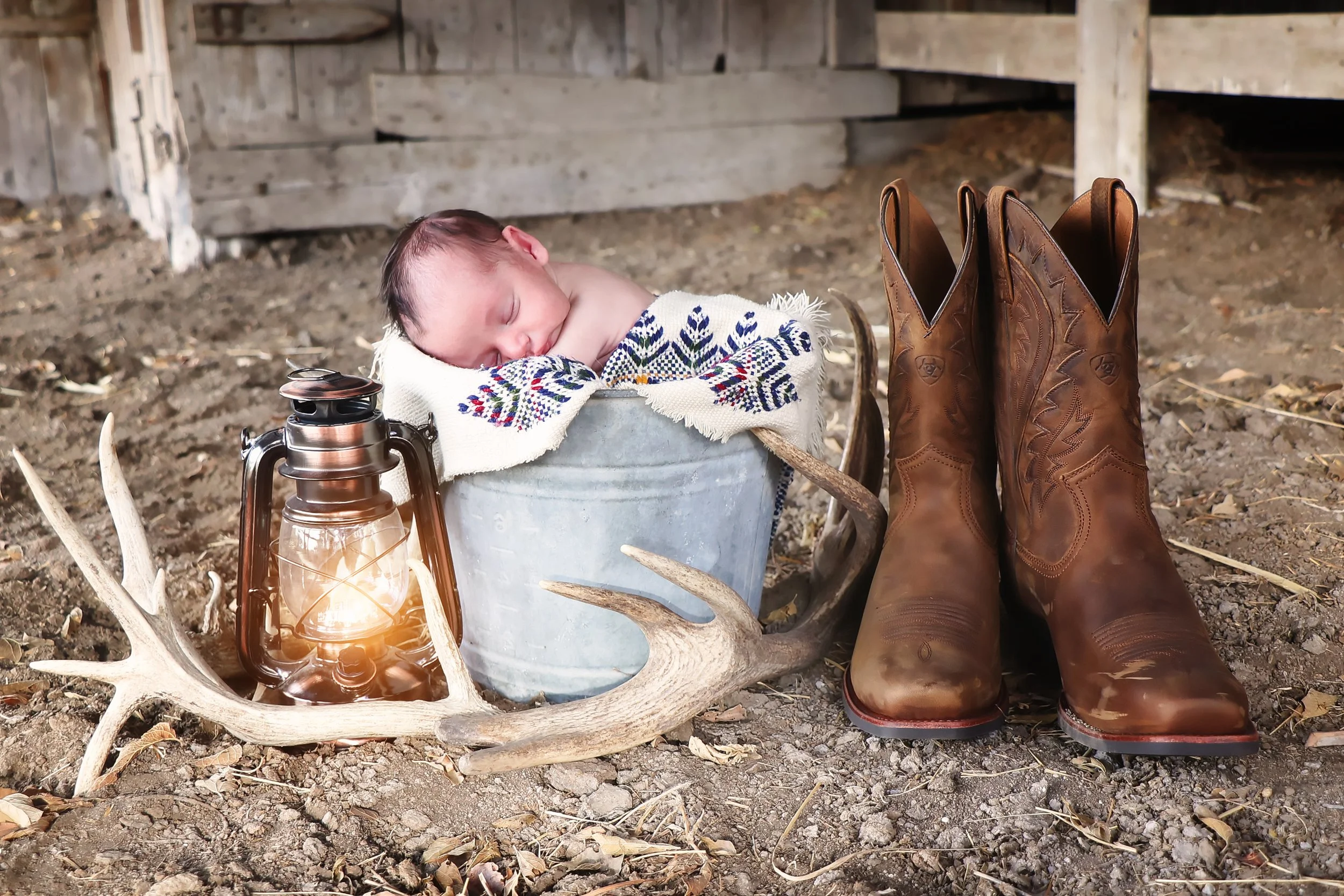 A sleeping newborn baby at a rustic setting in a metal bucket with a patterned cloth, nearby a lantern, antlers, and cowboy boots on dirt ground.