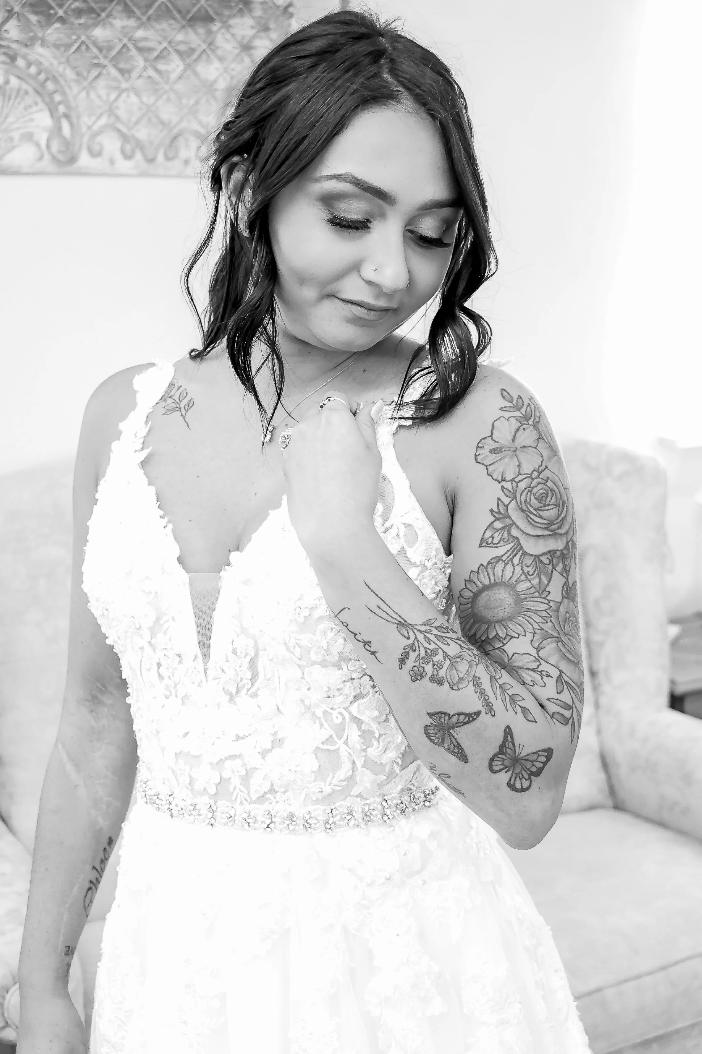 A woman in a white lace dress with dark hair and tattoos on her arm, looking down and holding her dress strap during her Bridal Photography session.