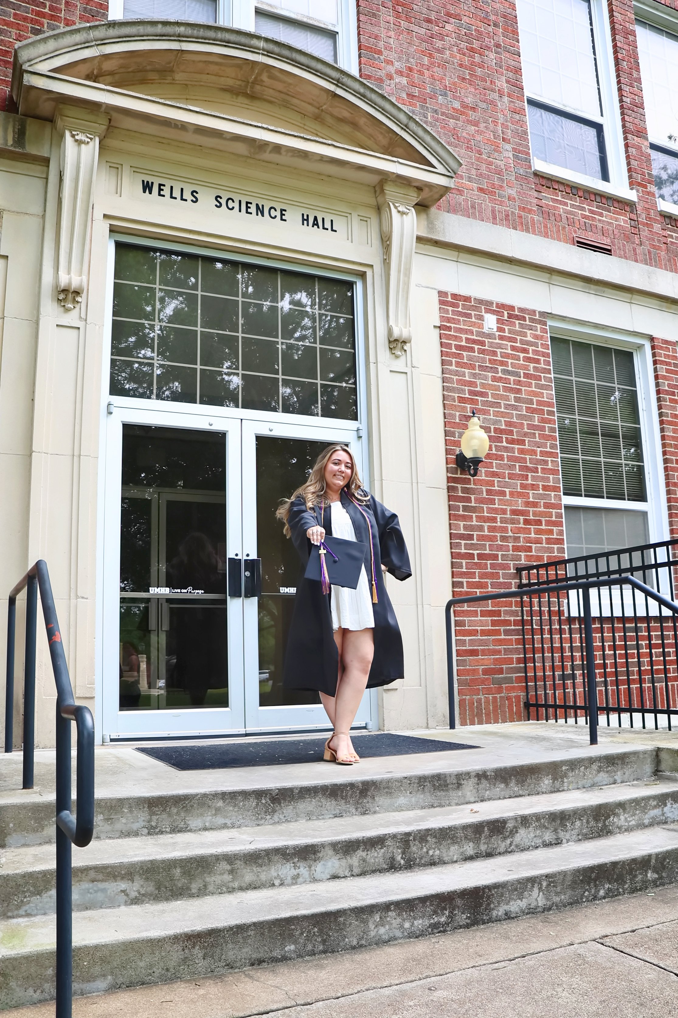 A woman in graduation attire standing outside the Wells Science Hall, holding a diploma and smiling at UMHB..