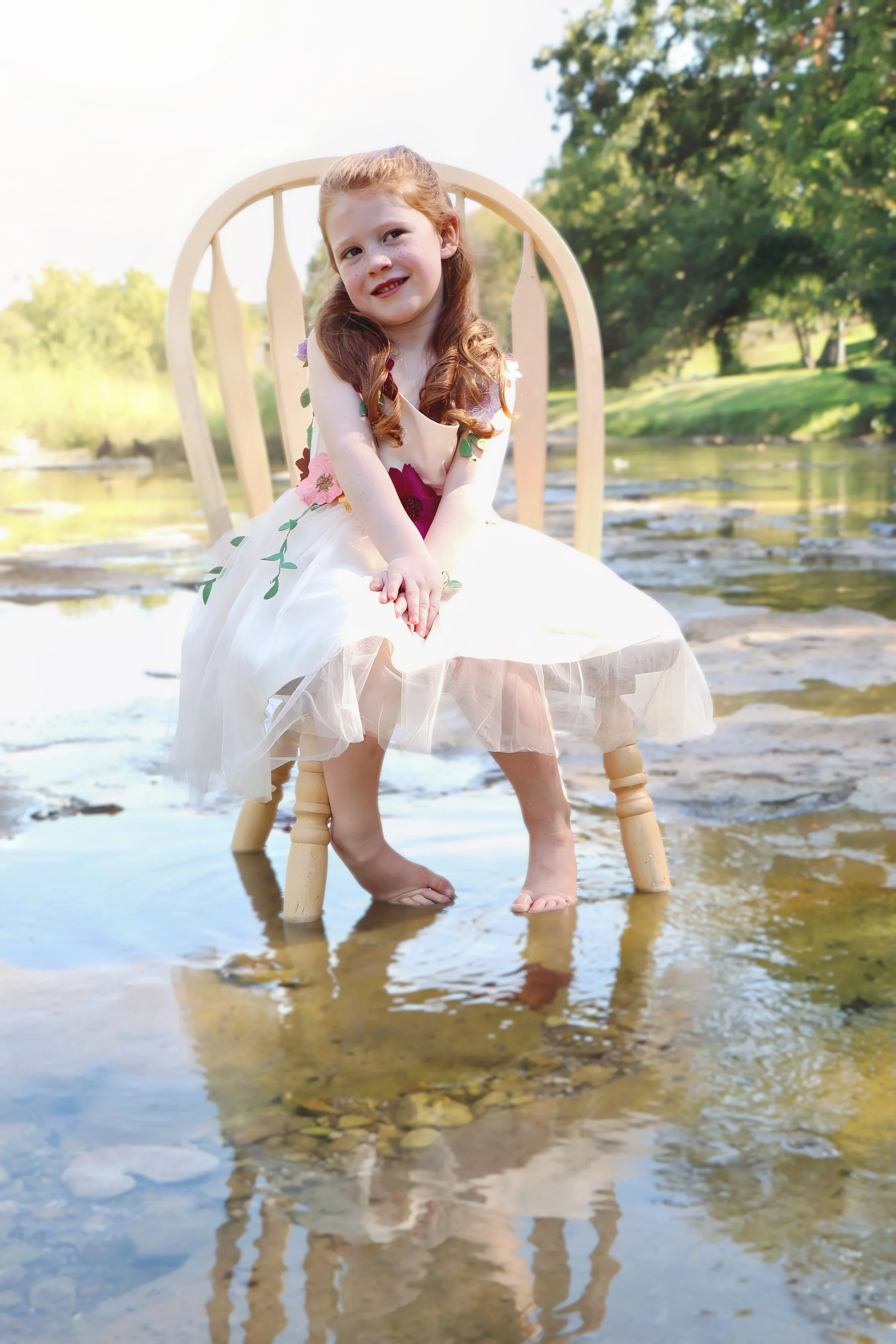 Young girl with red hair in a white dress sitting on a wooden chair in a shallow river, smiling, with trees in the background in Salado, Texas.