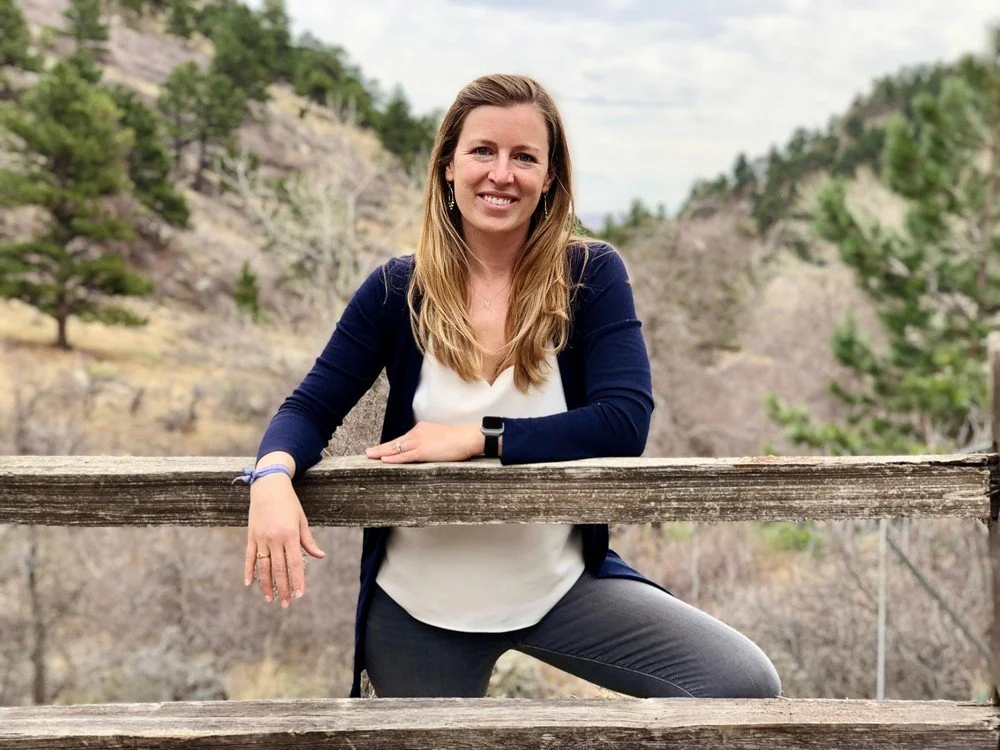 A woman with long light brown hair standing outdoors behind a wooden railing, smiling, with a scenic background of trees and hills.