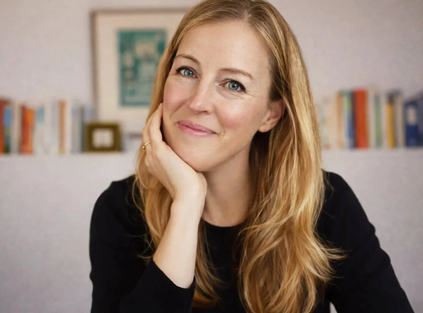 Portrait of a woman with long, wavy red hair, smiling gently, resting her face on her hand, in front of a blurred bookshelf background.