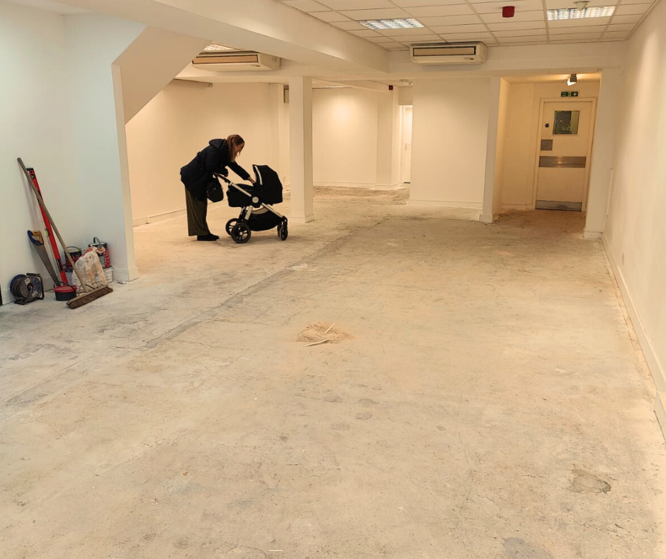 A woman in a black coat is pushing a pram in an empty, unfinished room with white walls and a concrete floor.