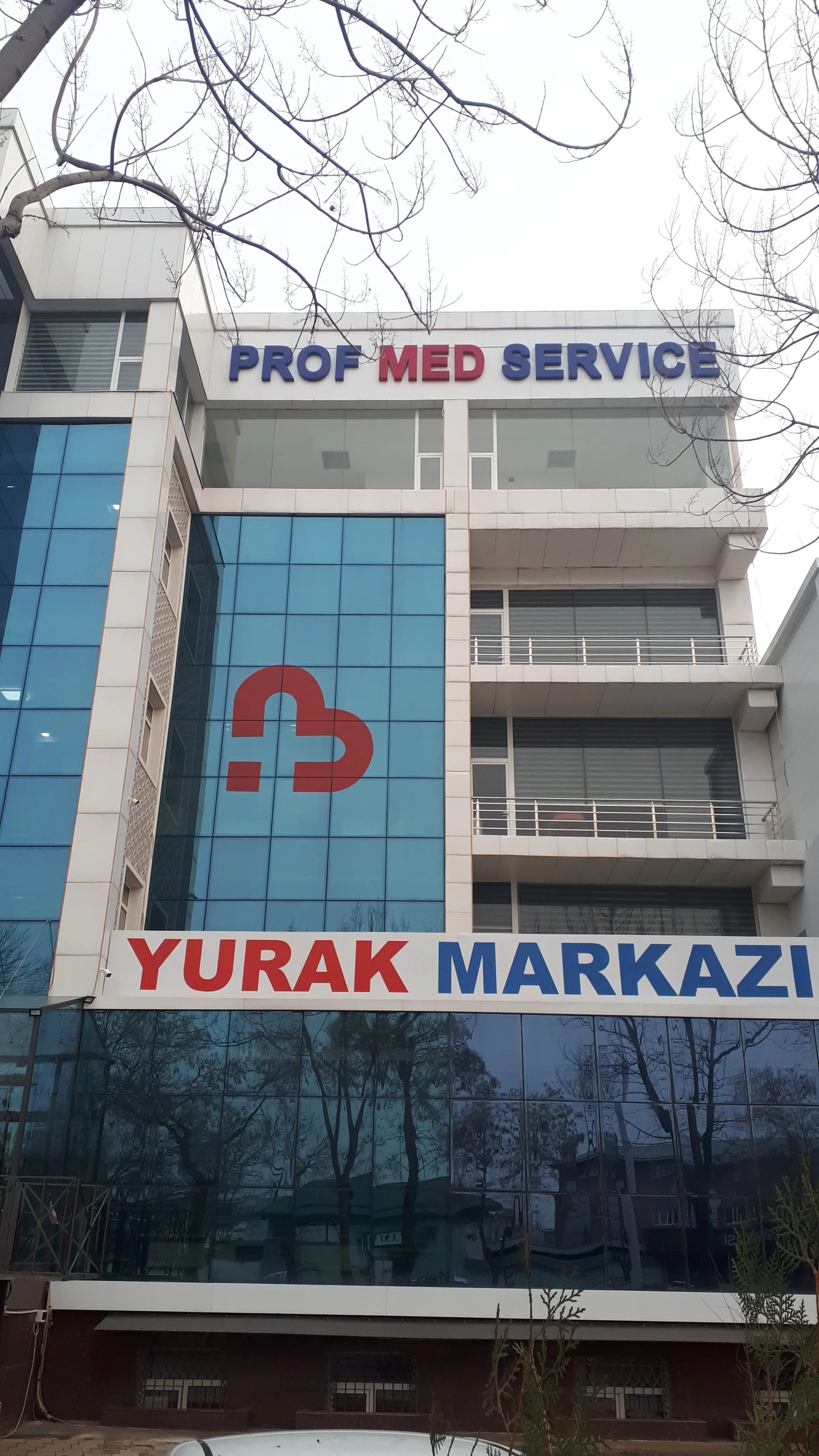 A multi-story medical building with signs reading "PROF MED SERVICE" at the top and "YURAK MARKAZI" on a banner below. The building has large tinted windows, and leafless tree branches are visible in the foreground.