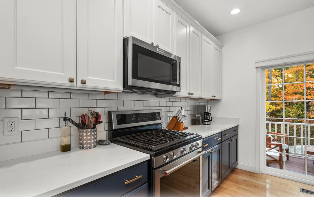 Modern kitchen with white cabinets, a stainless steel microwave, gas stove, and a sliding glass door revealing an outdoor deck with chairs and trees with fall foliage.
