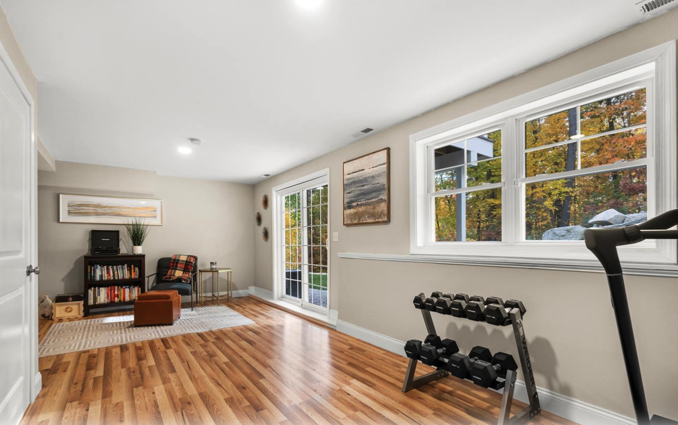 Living room with hardwood floors, large windows showing fall foliage outside, a small bookshelf, a chair with a blanket, artwork on the walls, and fitness equipment including dumbbells and a treadmill.