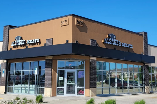 A  commercial building with a tan facade, black trim, and large glass windows. The sign reads "Alsager Meats" with a black silhouette of a cow above it. The building's address, 5675, is displayed on the upper corner.