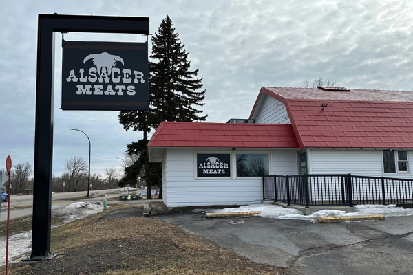 Exterior view of Alsager Meats storefront with a sign and a building with a red roof in a parking lot with some snow on the ground.
