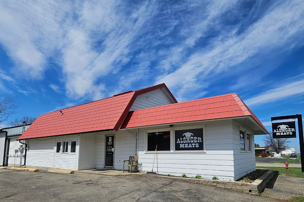 Single-story white building with a red roof, sign reading 'Alsager Meats', and parking lot in front, under a partly cloudy blue sky.