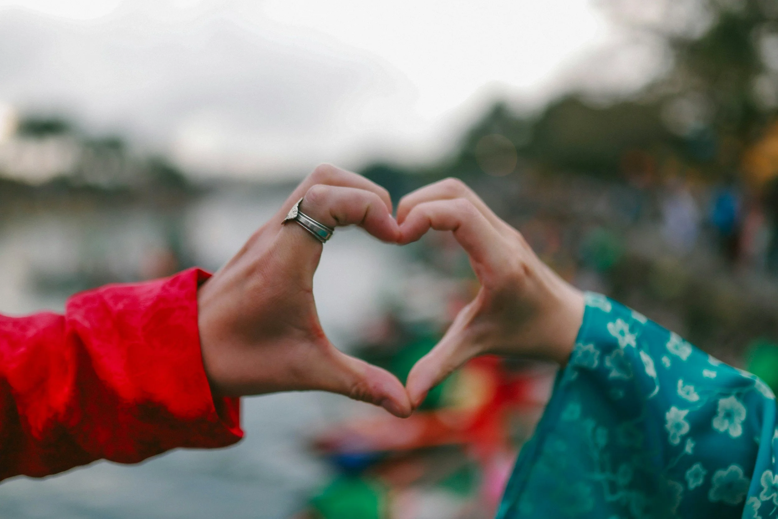 Two people in colorful jackets form a heart shape with their hands, with a blurred outdoor background.