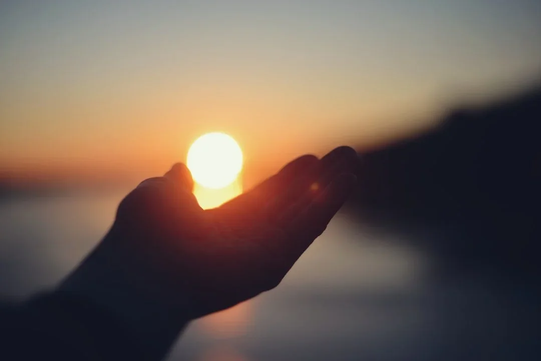 A hand holding the setting sun during sunset, with hills or mountains in the background.