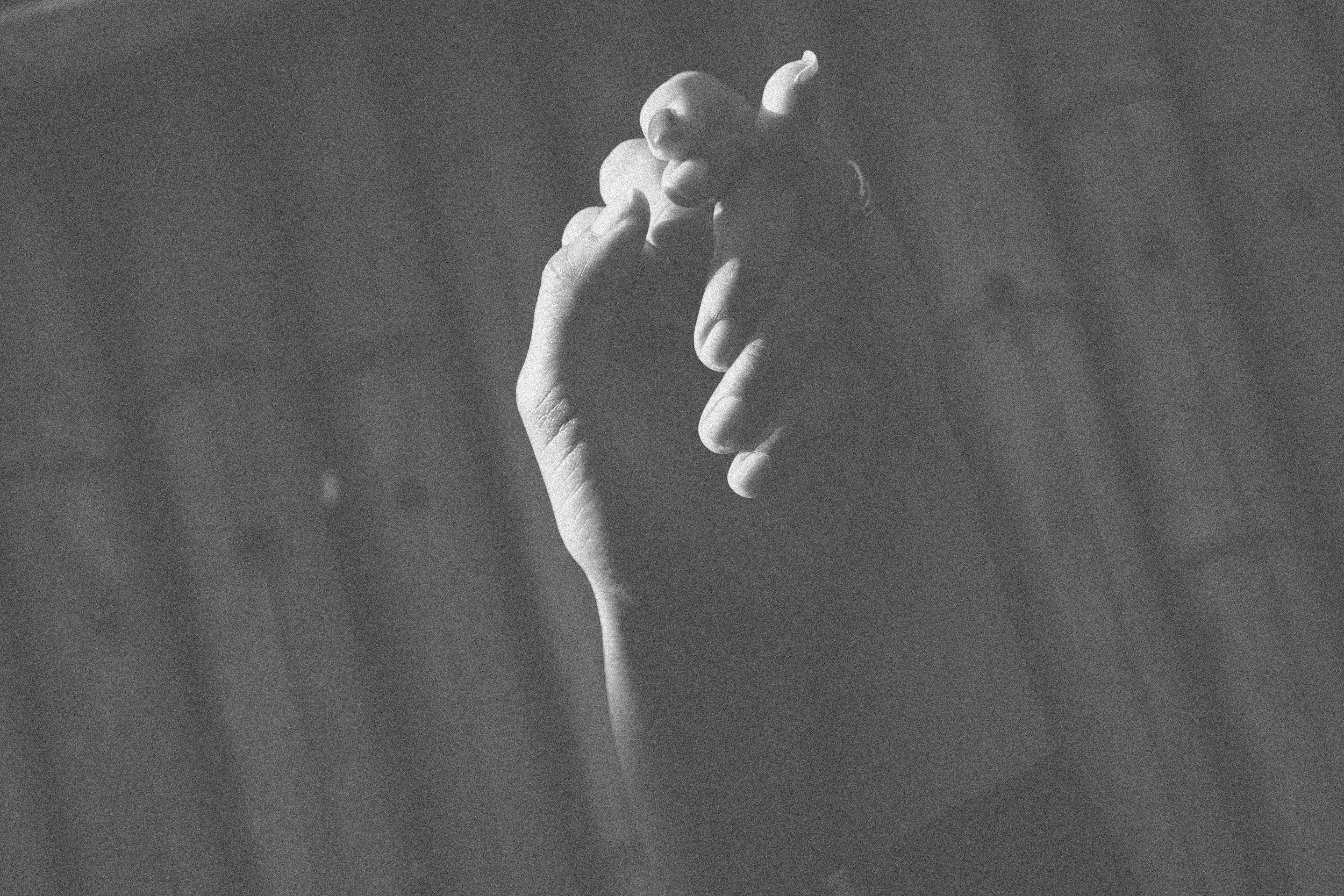 A black-and-white photograph of a hand with fingers intertwined and visible nails, positioned against a dark background.