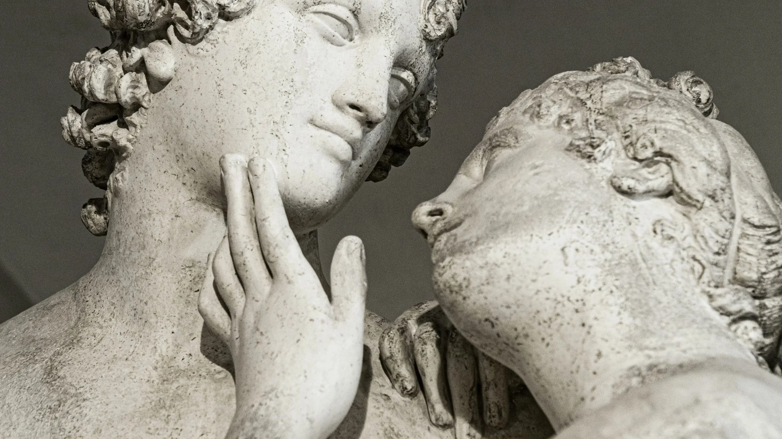 Close-up of two classical sculptures depicting a man and a woman in a close, intimate pose, with the woman gently touching her lips with her fingers.