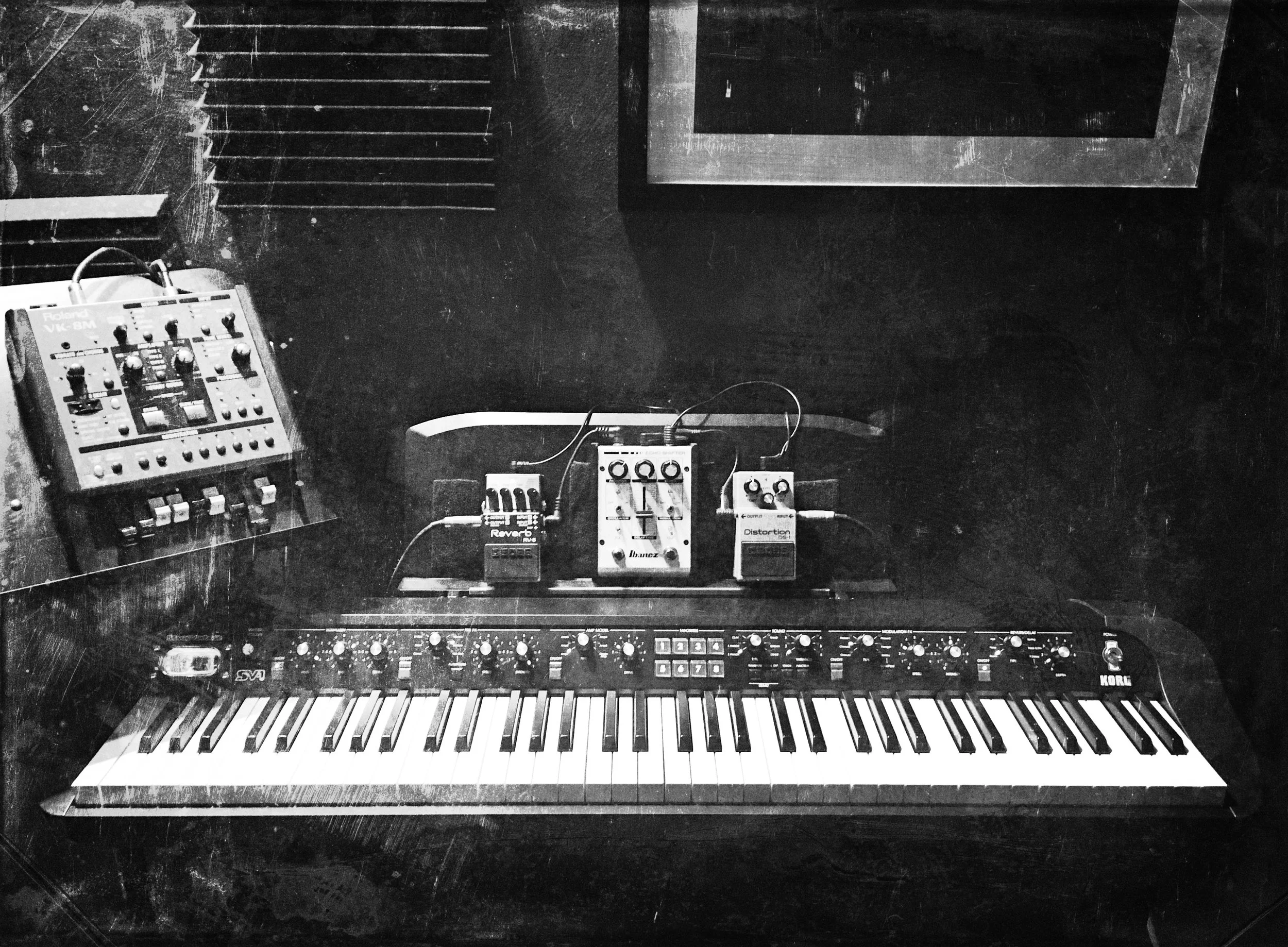 Black and white photo of a music studio setup featuring a Korg digital piano running through guitar pedals. To the left is a Roland VK-8M organ unit. 