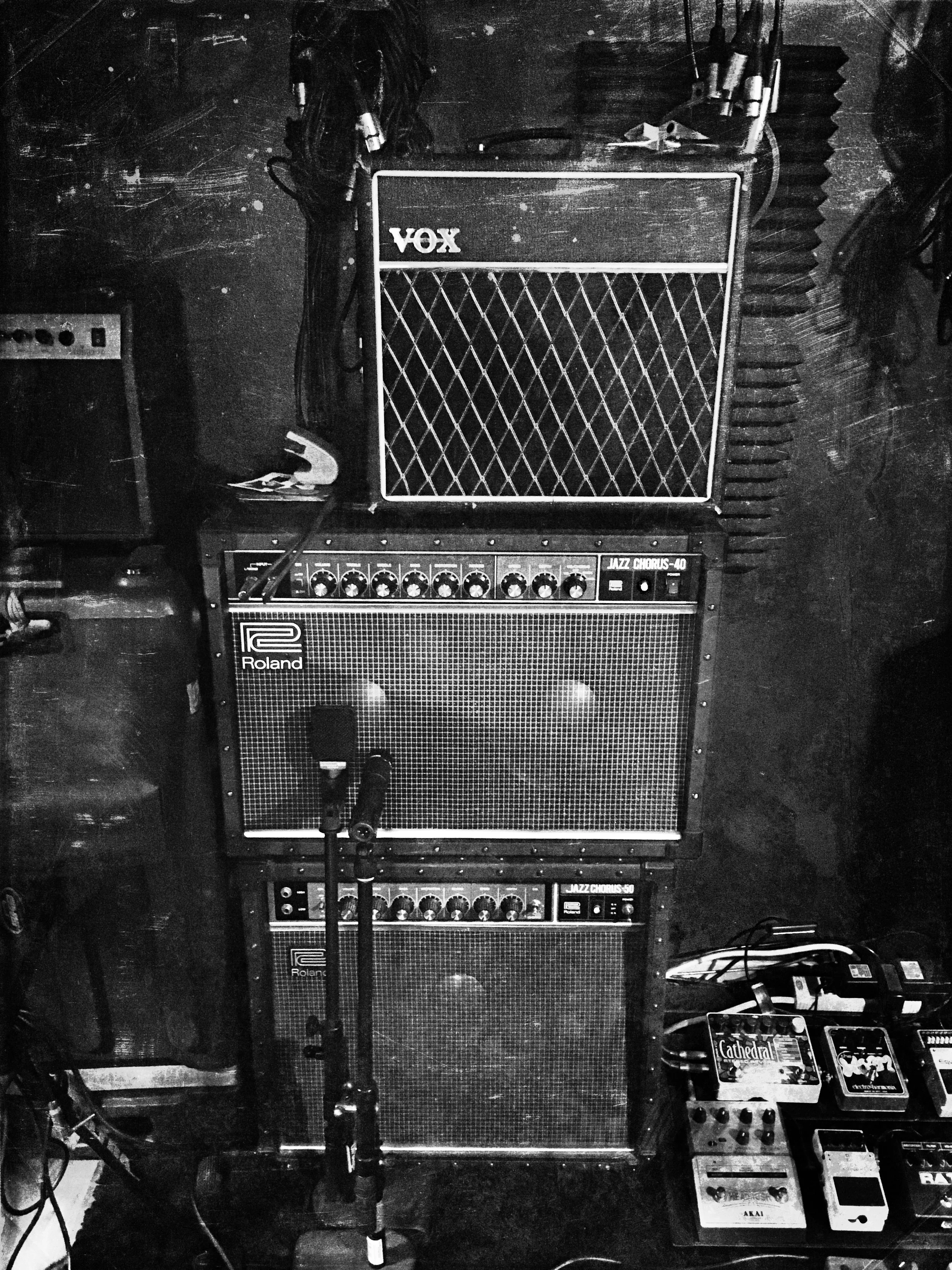 A black and white photo of musical equipment, including an amplifier stack with two Roland Jazz Chorus guitar amplifiers and a VOX amplifier on top, with various effect pedals and an electric guitar nearby.