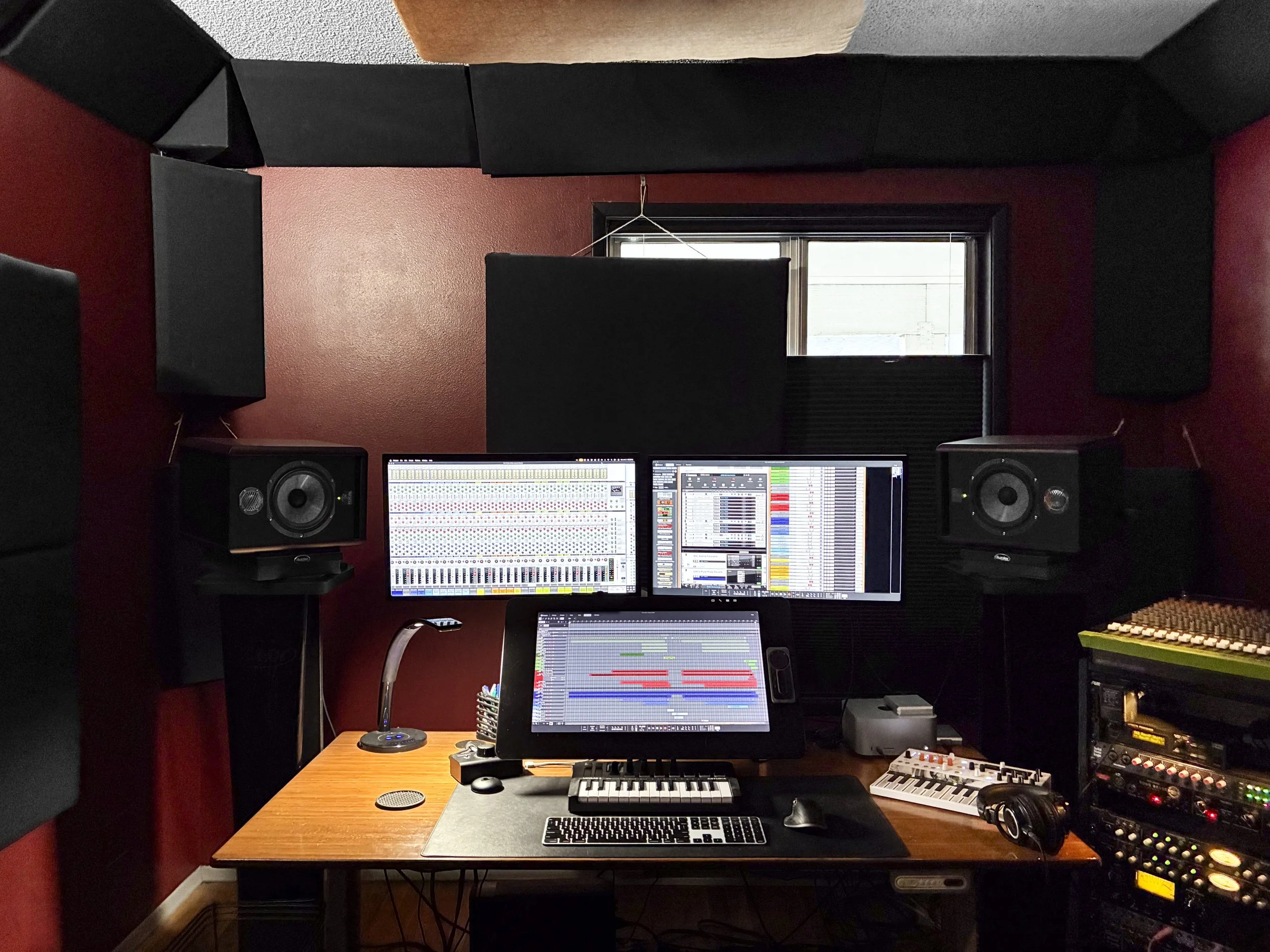 A music production studio with a desk holding a computer with three monitors, a MIDI controller, a keyboard, and a mouse. Two studio monitors are on either side of the monitors. The studio is underwater research and design, Jeremy Wilkins' studio. 