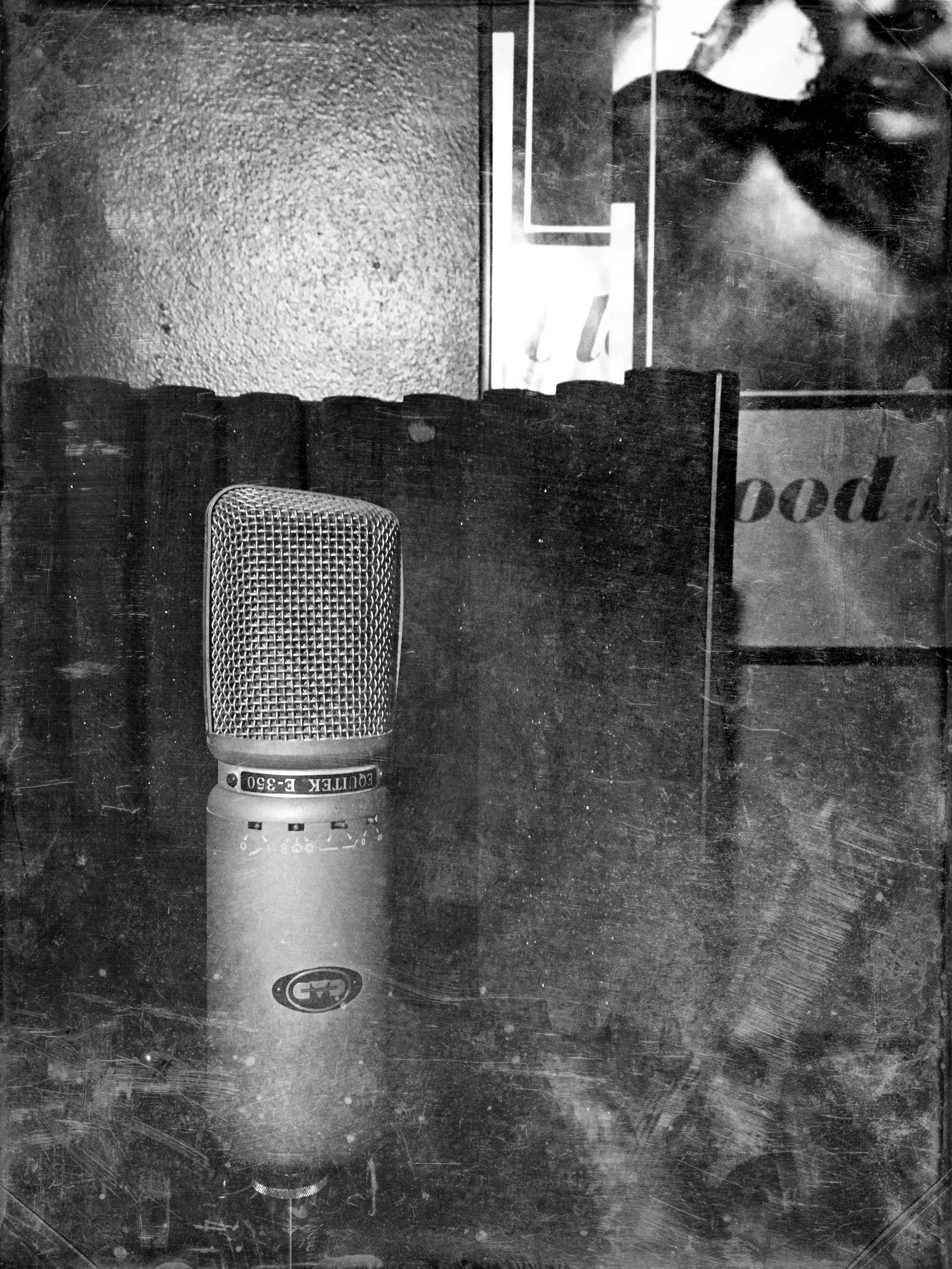 Black and white photo of a condenser microphone on a stand in a recording studio, with a textured wall and a partial view of a poster for the album, "Blood" by This Mortal Coil in the background.
