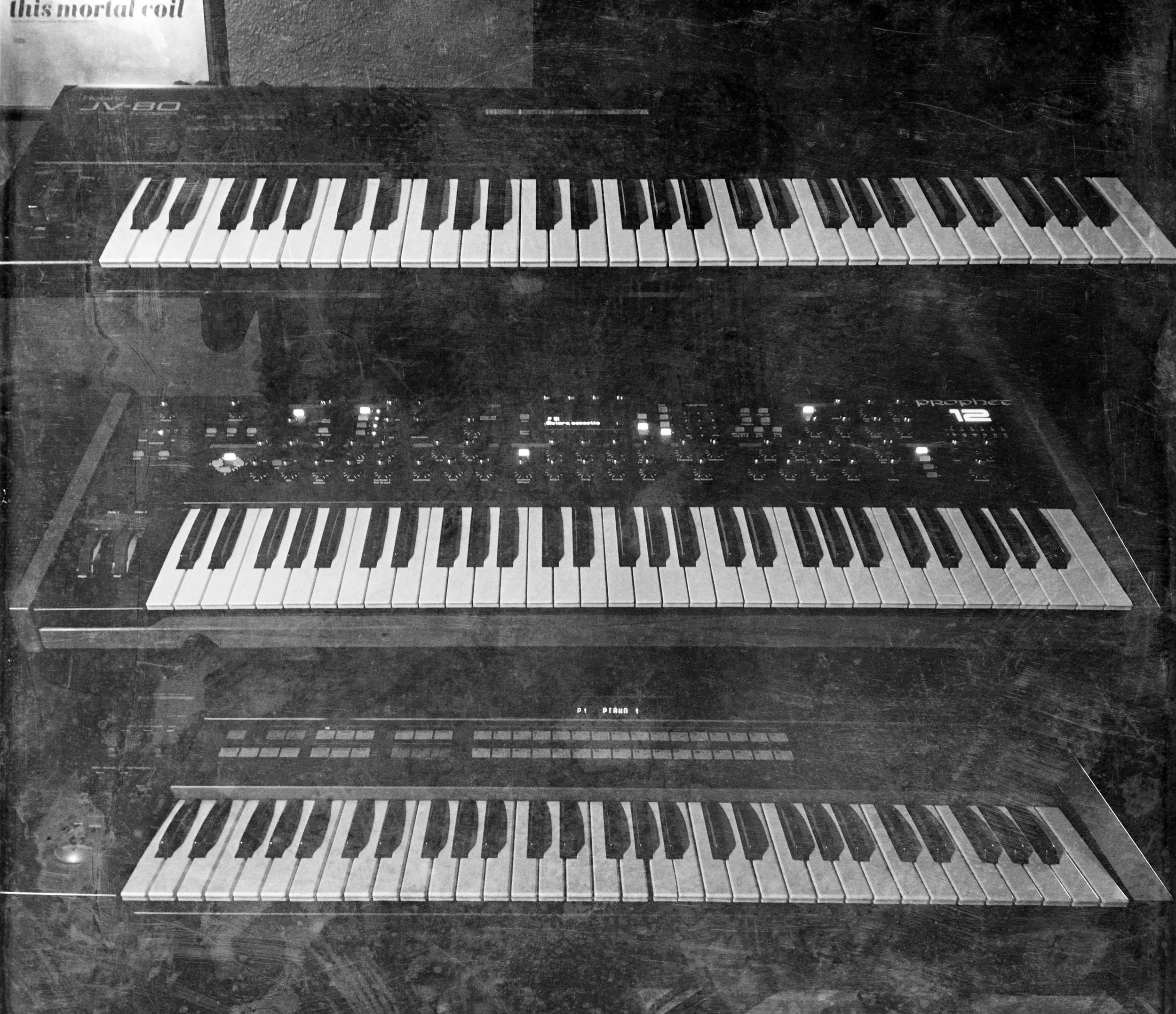 A black and white photo of three electronic musical keyboard synthesizers stacked vertically, including a Roland JX-8P, a Prophet 12, and a Roland JV-80, with visible controls and keys.
