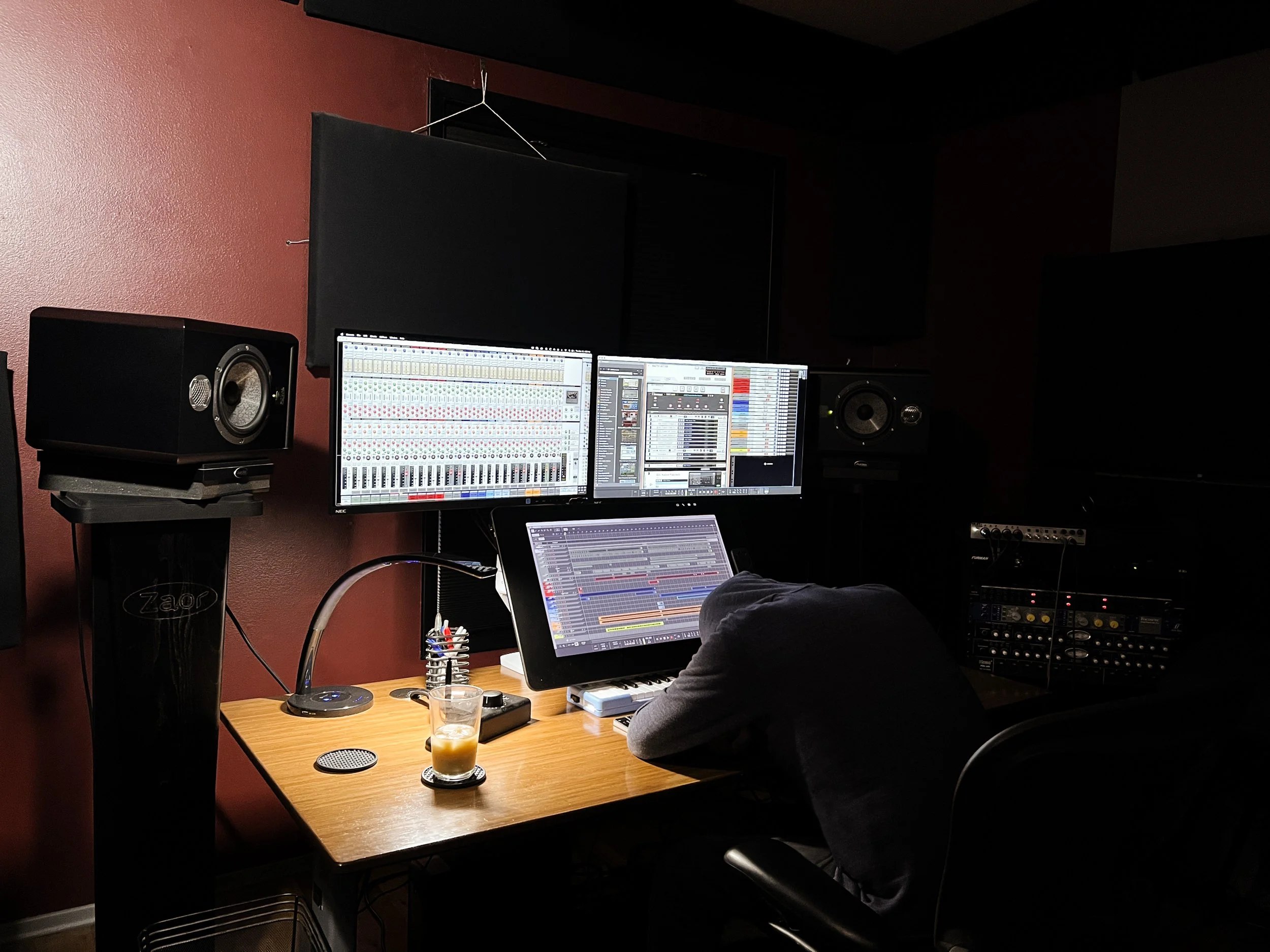 Jeremy Wilkins, a music producer working at underwater research and design. The desk has multiple monitors displaying Reason, studio speakers, and audio equipment, with a glass of iced coffee on the desk. 