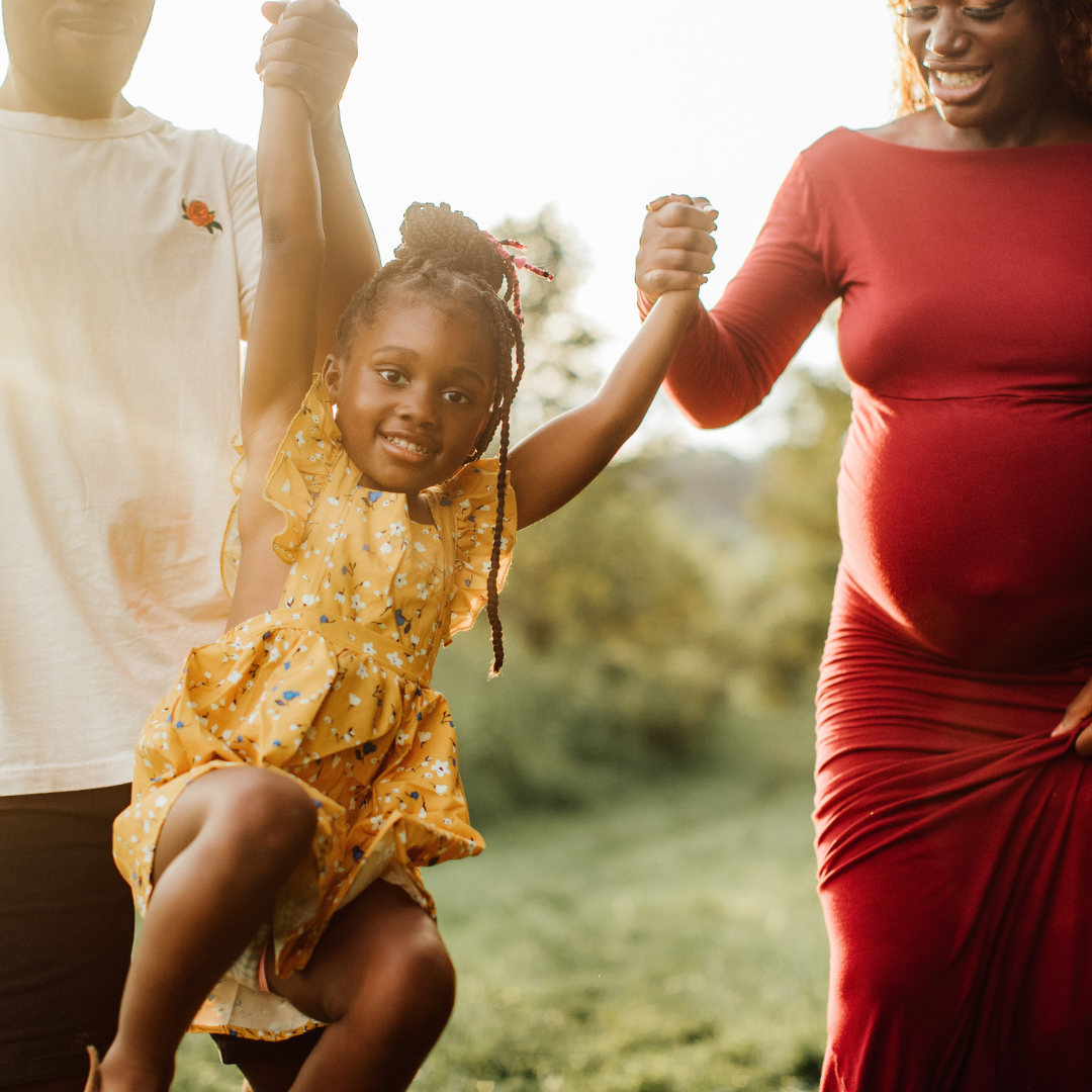 A young girl in a yellow dress is swinging and smiling while holding hands with two adults, one on each side, outdoors in a park or garden during daytime.