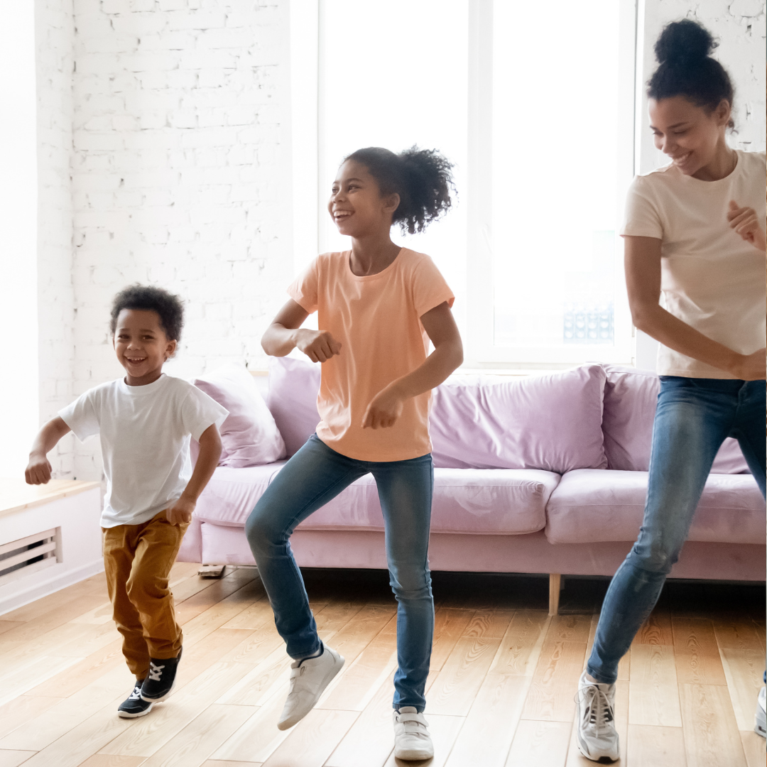 Three children dancing joyfully in a living room with a white brick wall and a pink sofa in the background.