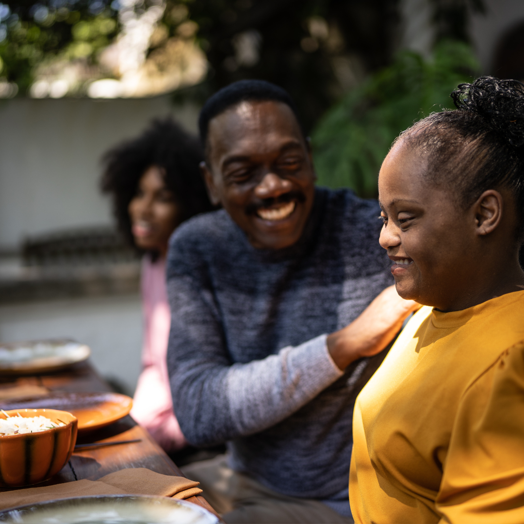 Three people smiling and enjoying each other's company at an outdoor meal table, with bowls of food visible.