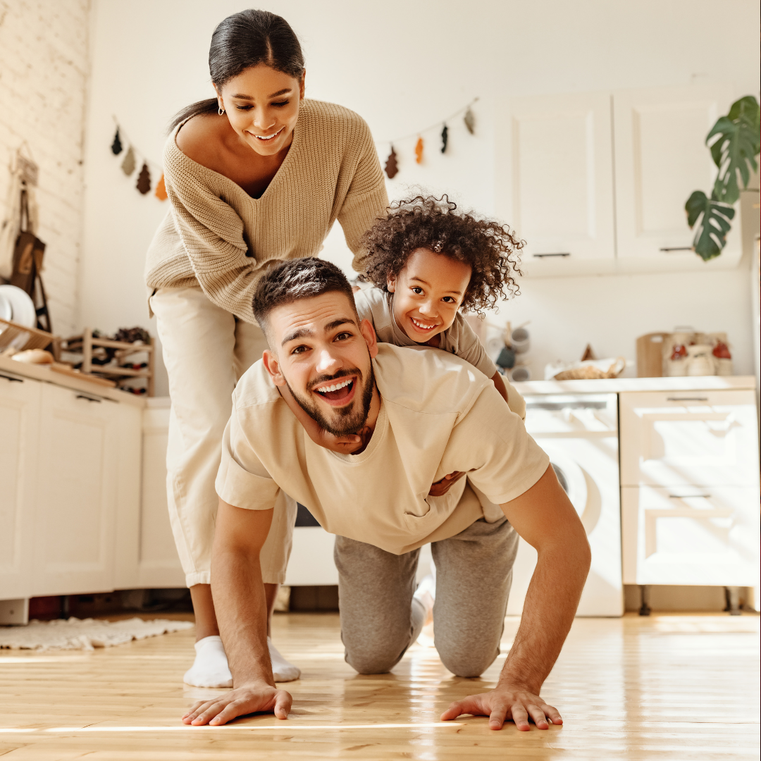 A happy family of three playing on the floor of a bright kitchen, with the woman on top, the man in a crawling position, and their daughter between them, all smiling.