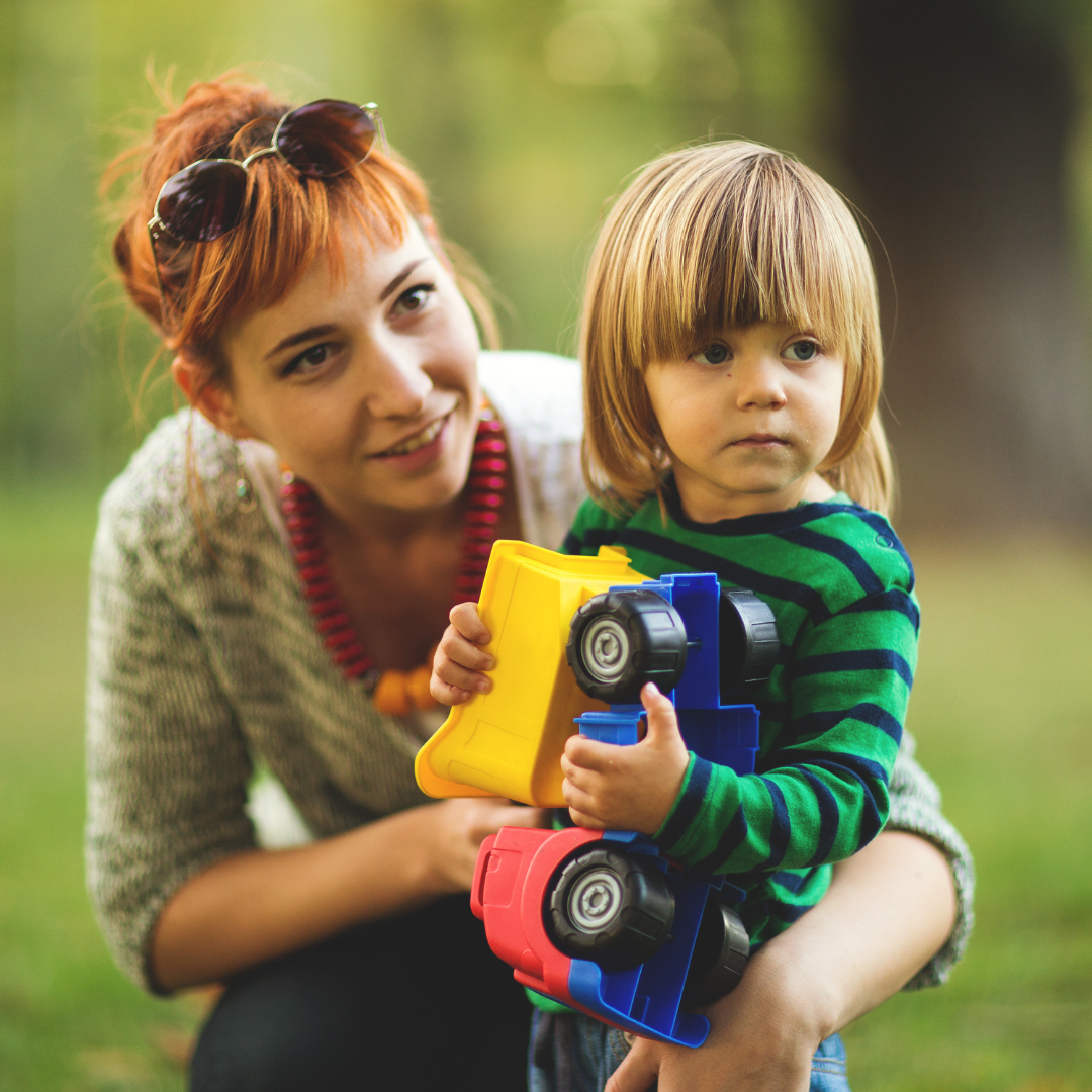 A young boy with straight, shoulder-length blonde hair holding colorful toy trucks while standing outdoors, with a woman with short red hair, sunglasses on her head, crouching beside him, outdoors in a park.