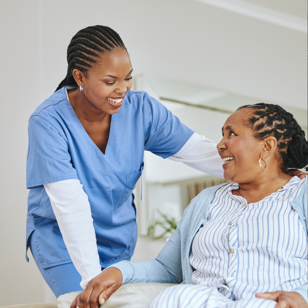 A healthcare professional, wearing blue scrubs, smiling and talking with an elderly woman in a hospital gown, seated in a hospital room.