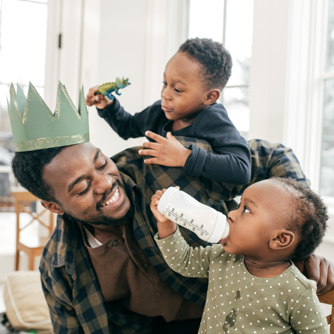 A man wearing a green paper crown, smiling with two children, one holding a toy dinosaur and the other drinking milk from a bottle, in a bright room with large windows.